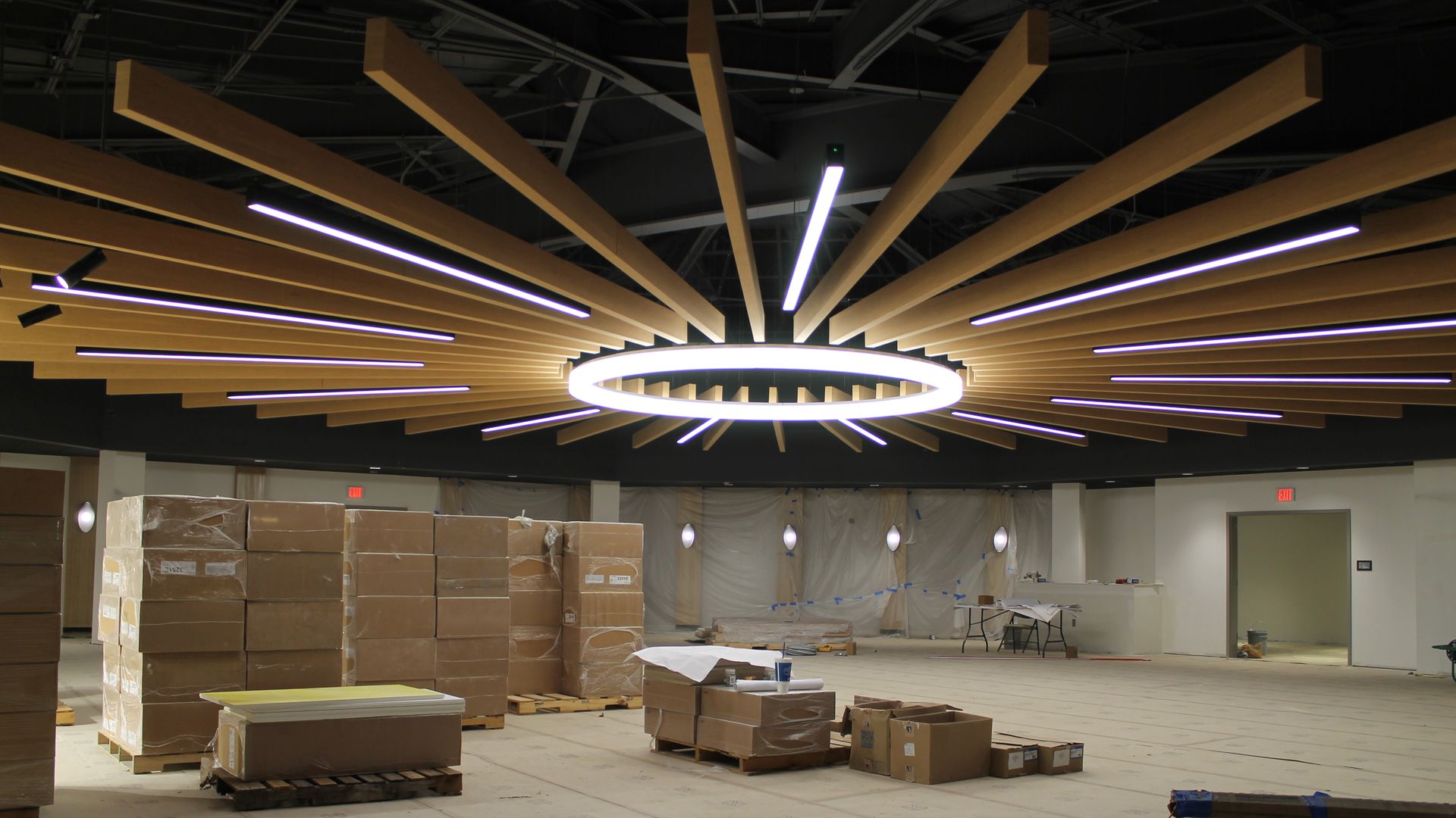 Interior construction site with a sunburst ceiling of wooden slats around a glowing circular ring light; purple LED strips radiate outward. Stacks of cardboard boxes and pallets on the floor.