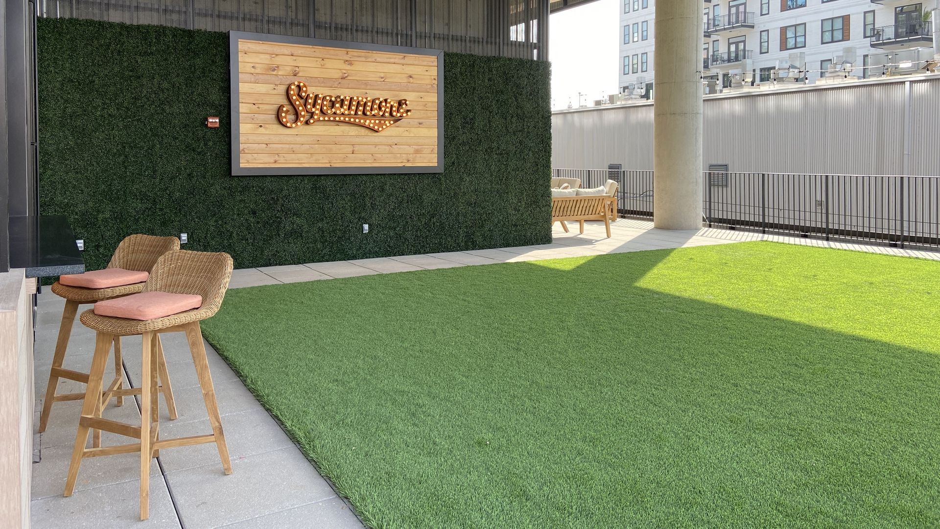Outdoor seating area with two wooden stools with pink cushions, artificial grass, a green hedge wall, and a wooden sign lit up spelling "Sycamore" under a concrete ceiling near an apartment building.