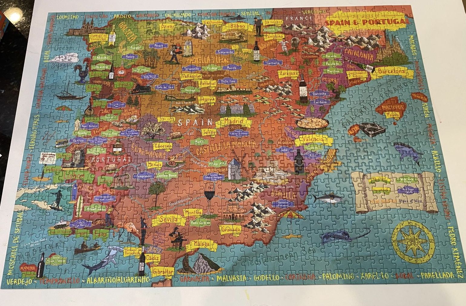 Completed colorful puzzle map of Spain and Portugal highlighting cities, wine regions, bottles, cultural icons, surrounding sea with fish, and mountains in the northern region.