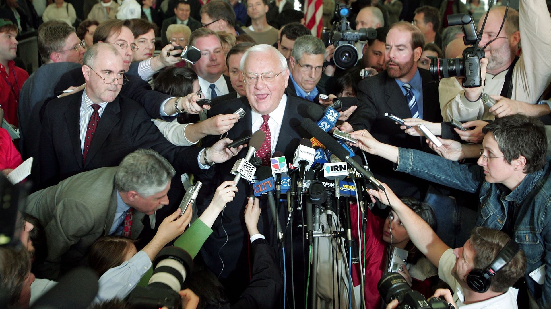 Photo of a man speaking into several microphones held by reporters.