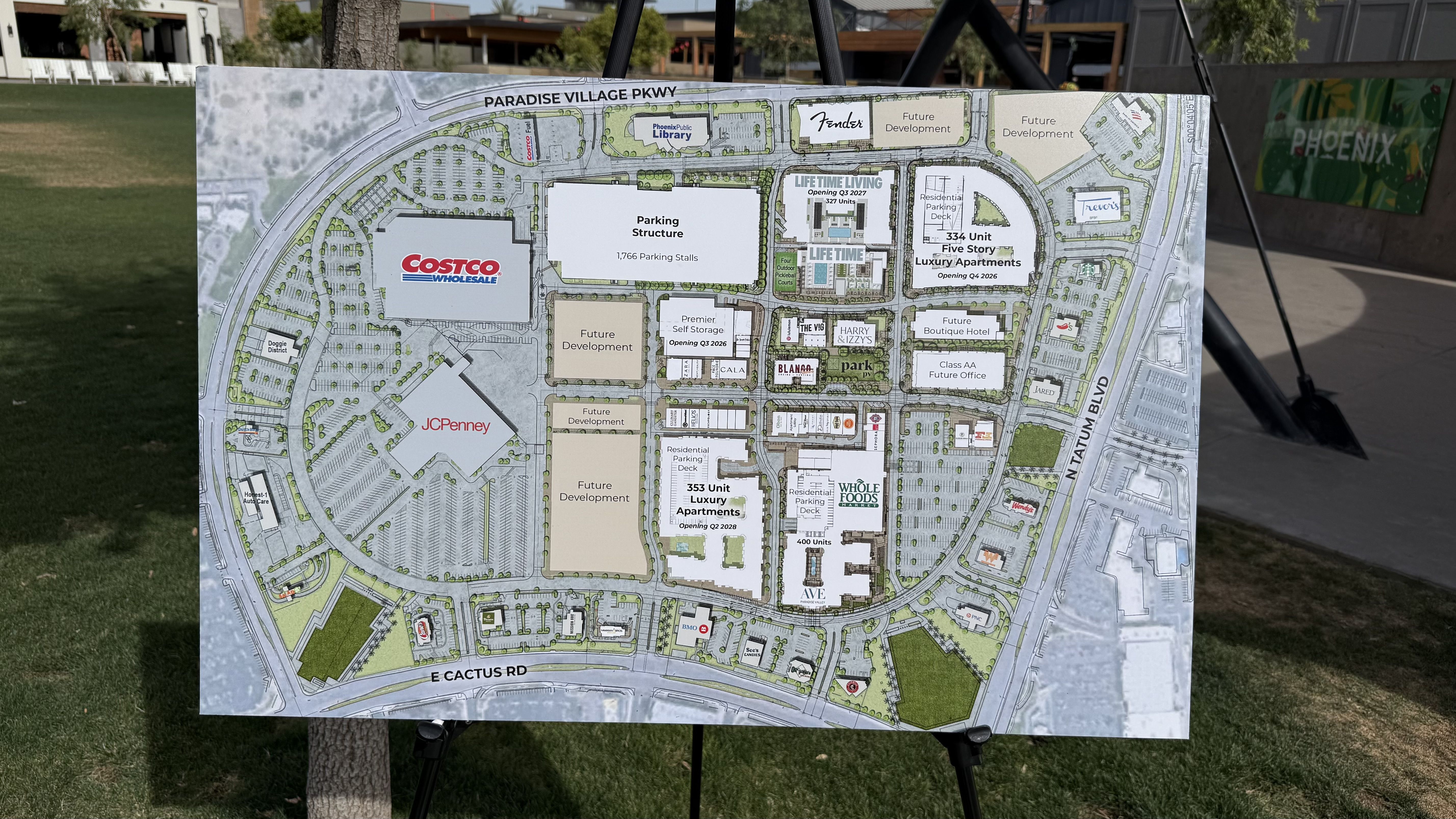 Large site map showing retail complex plan with Costco and JC Penney buildings, parking structures, and future development areas; mounted on a stand outdoors near a tree on a grassy area.