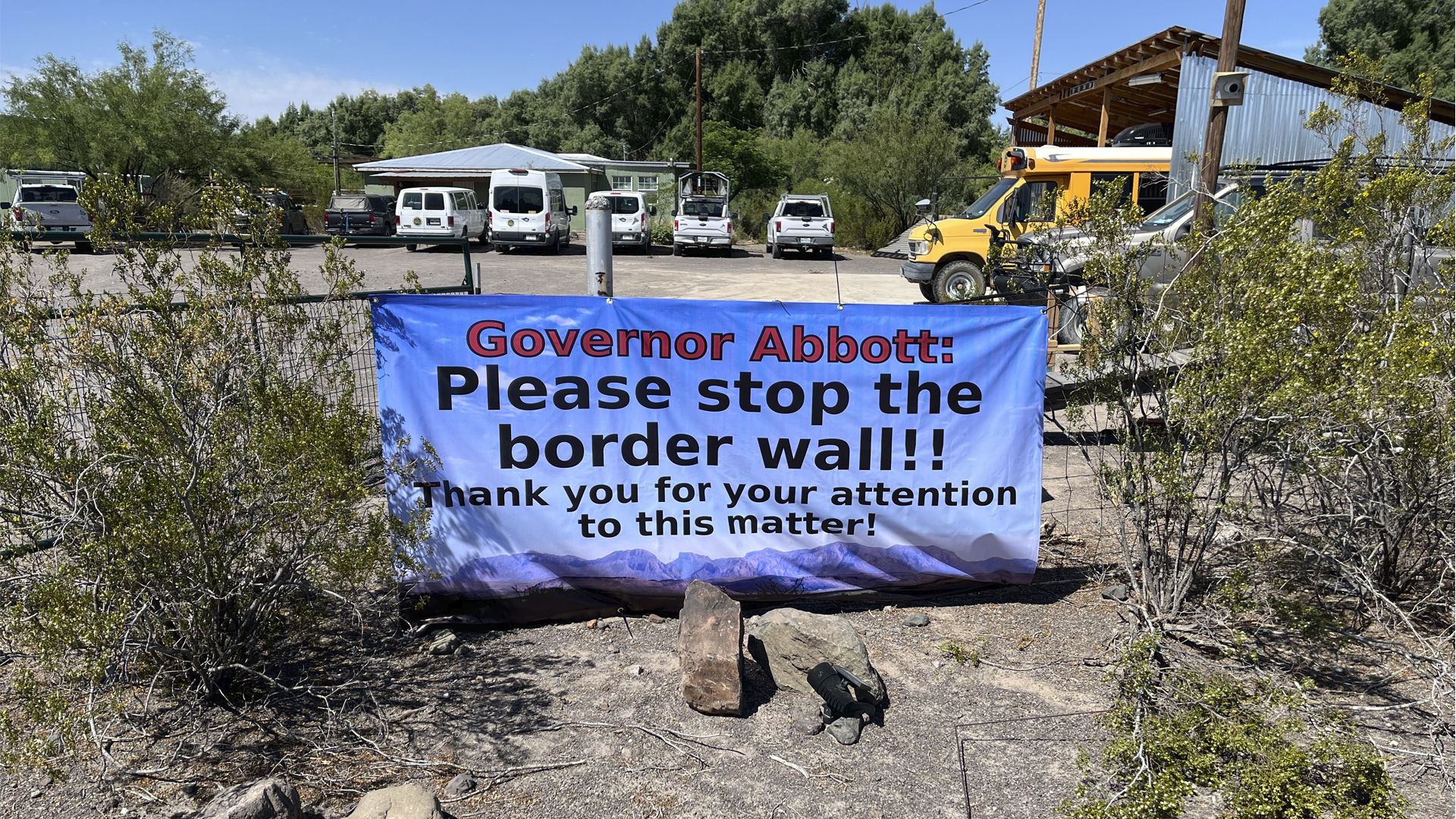 Blue banner reading "Governor Abbott: Please stop the border wall" hangs on a fence above a gravel lot with white vans, a yellow school bus, and a metal shed, under clear blue skies.