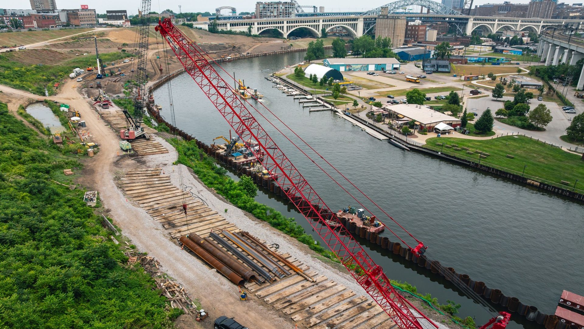A red crane and construction equipment on a riverbank with steel pipes and wooden planks, near a waterway with docks and urban bridges in the background under overcast sky.
