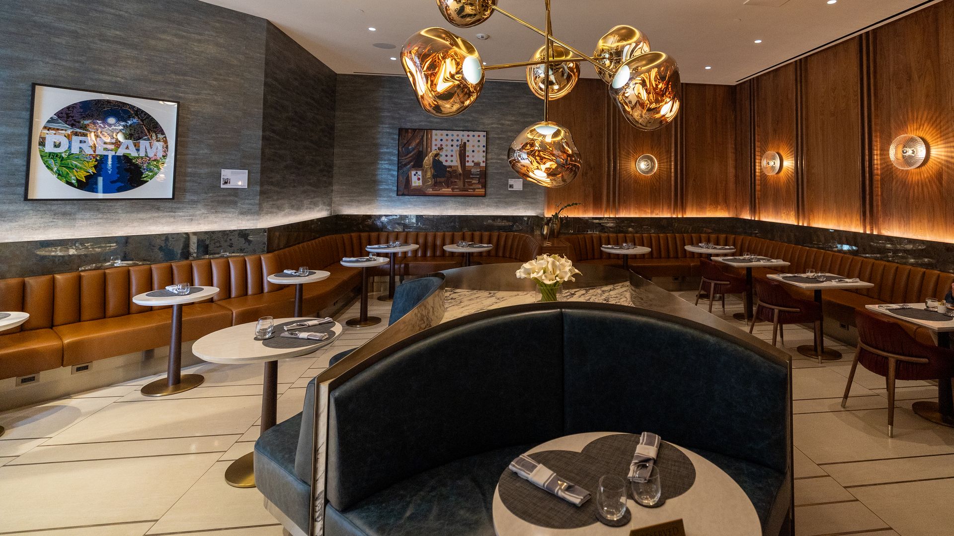 Elegant restaurant interior with a curved navy banquette in the foreground, round marble tables with napkins and glasses, a large gold orb chandelier, wood walls, and framed art featuring DREAM
