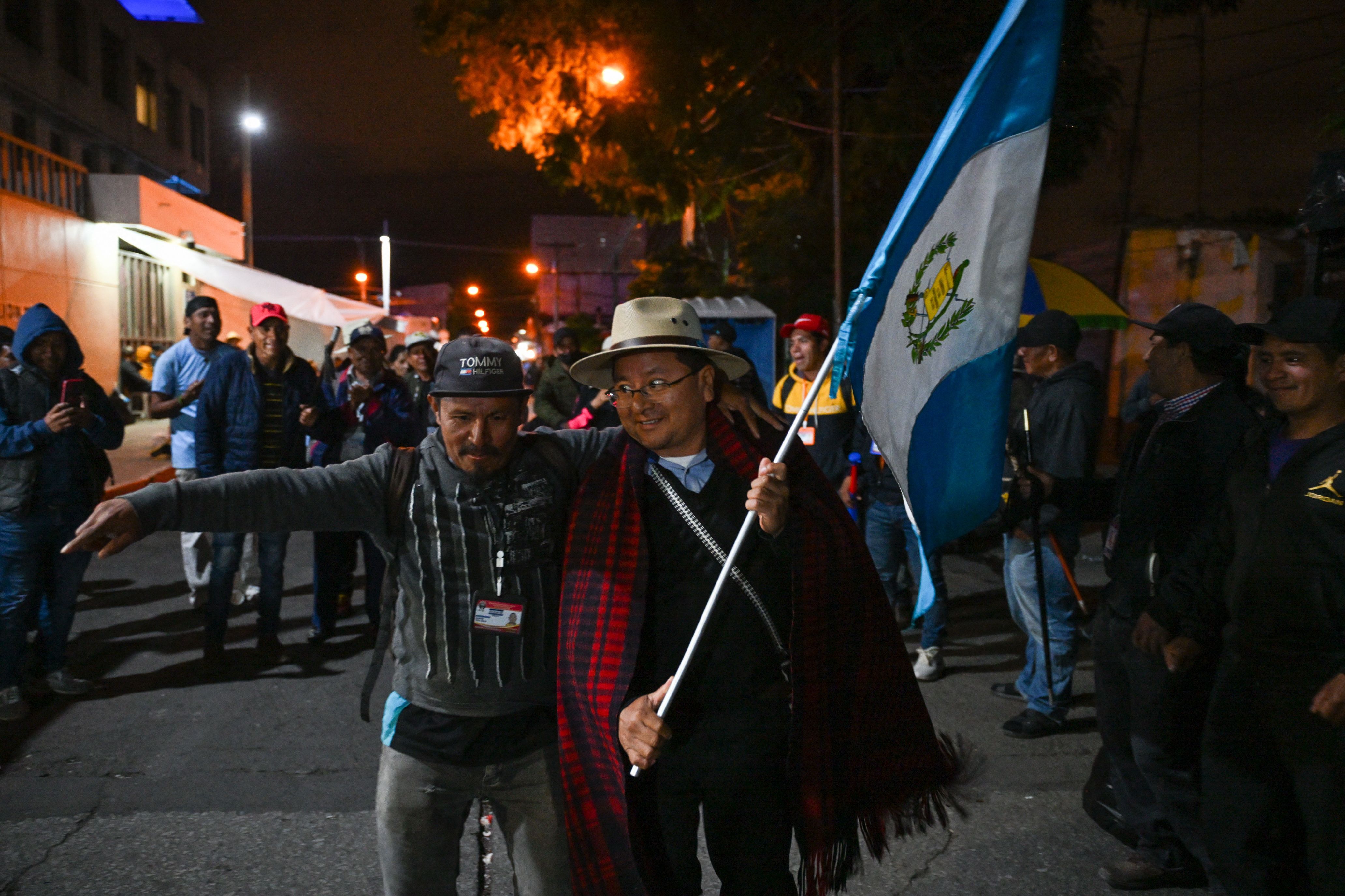 two Guatemalan men walk side by side, one holding a large Guatemalan flag and the other with one arm in the air. 