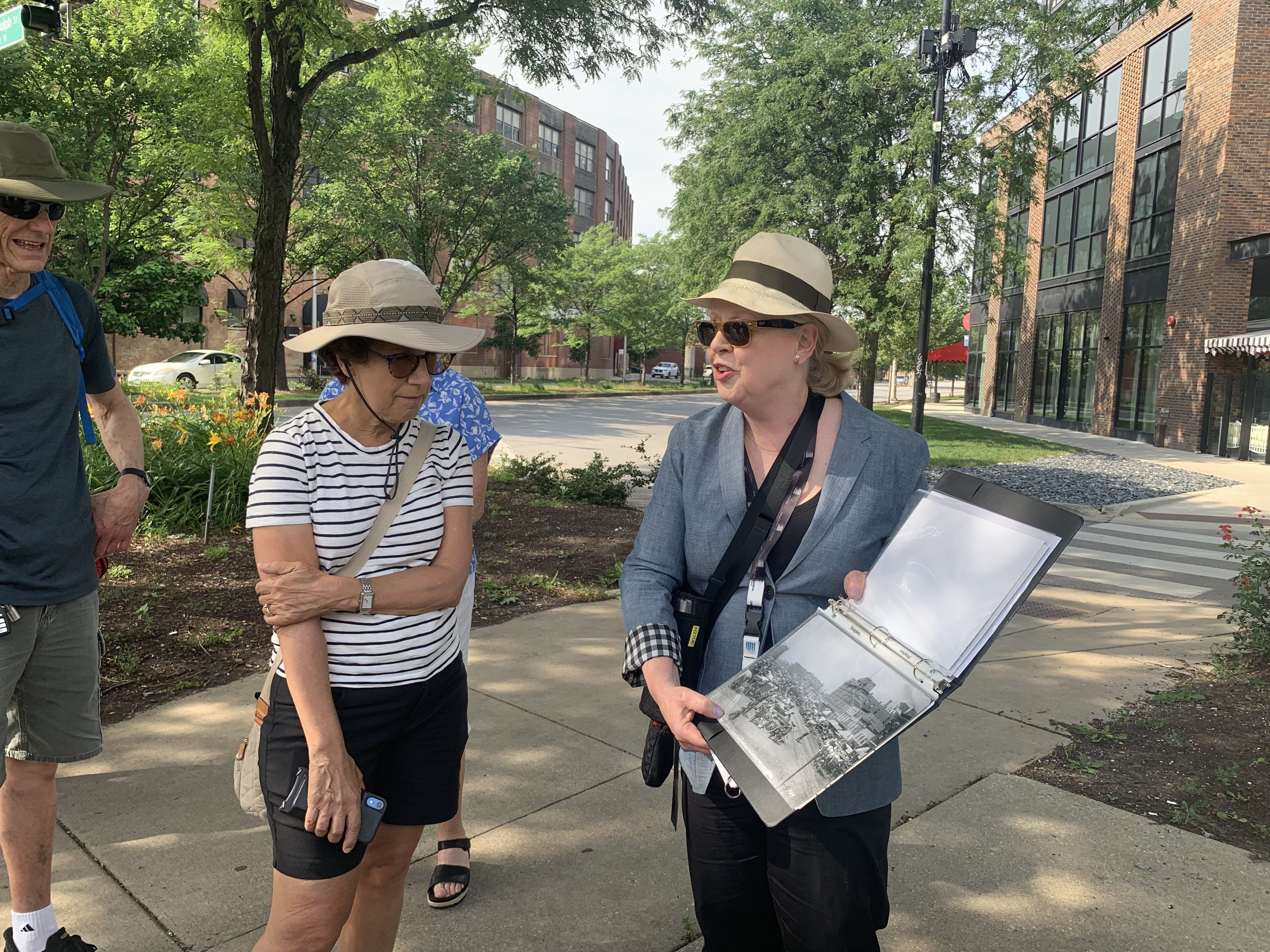 A woman in a gray blazer and beige hat stands outdoors holding a binder with black-and-white photos, talking to a woman in a striped shirt and hat, with trees and buildings in the background.