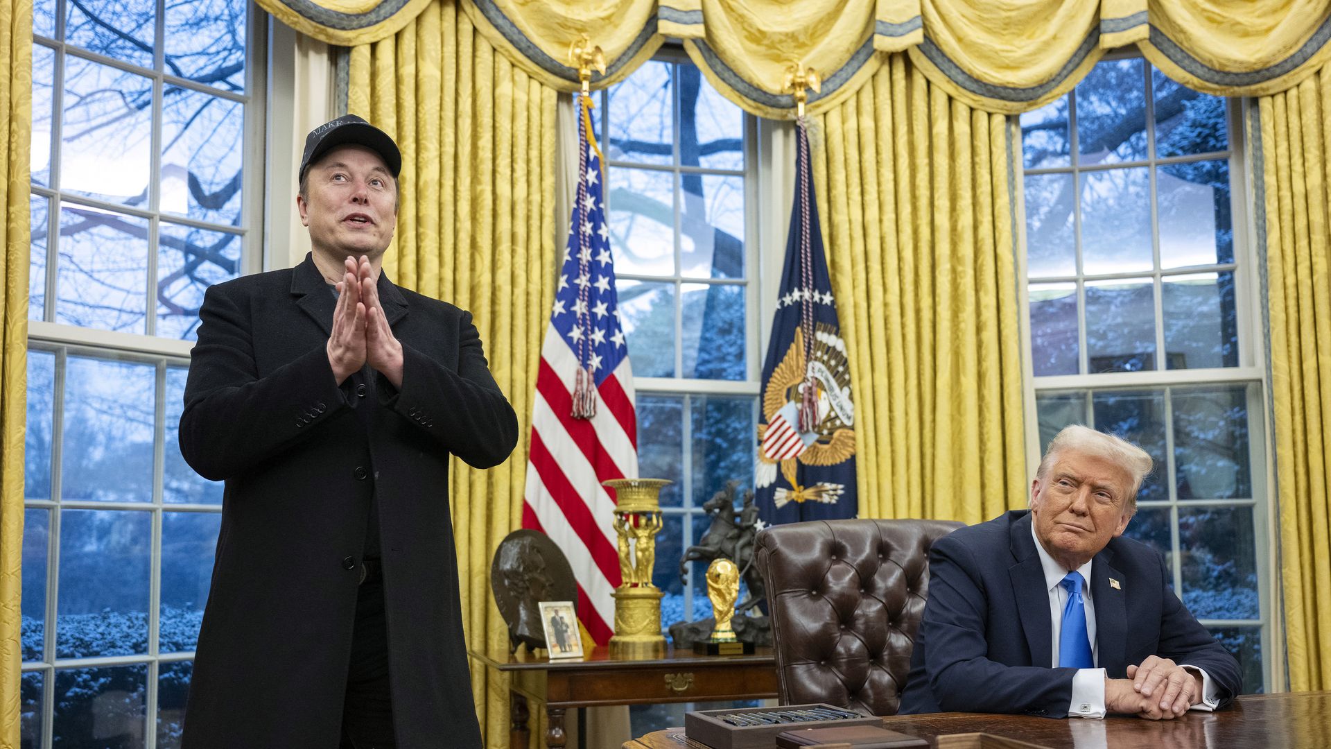 Elon Musk stands in the Oval Office while President Trump, seated at his desk, looks on.