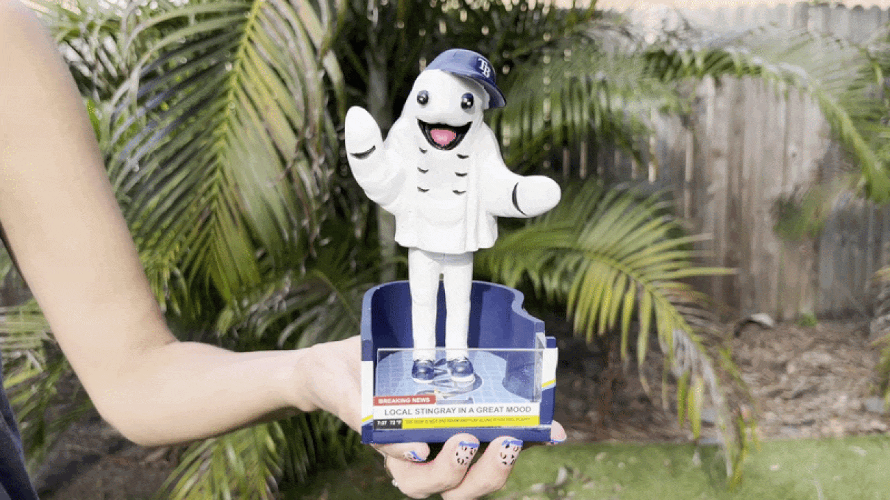 White cartoon manatee wearing a blue baseball cap with open arms, perched on a clear base held by a hand in a tropical garden; a blue display box sits beneath.