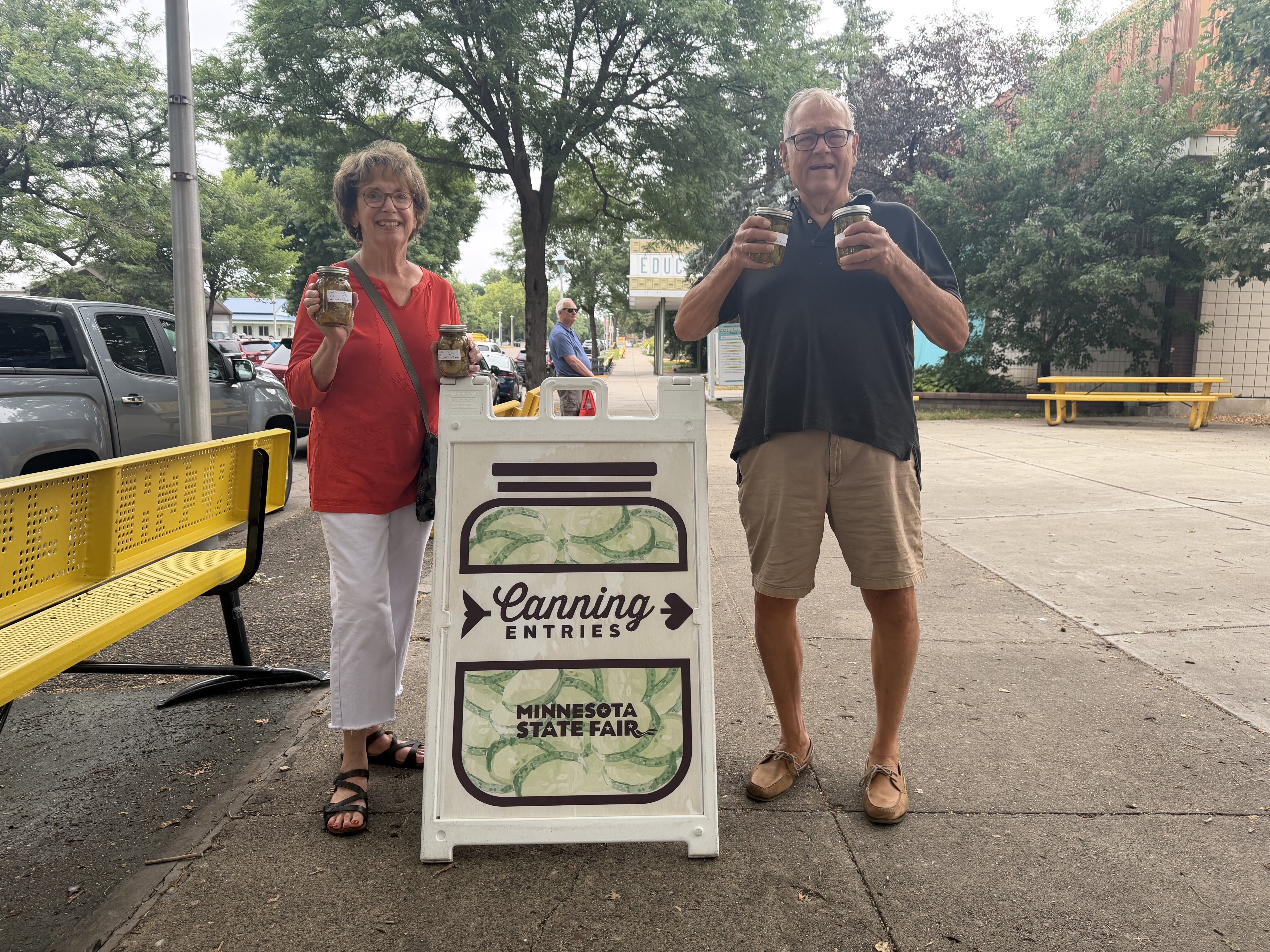 Two smiling seniors hold jars of canned goods on a sidewalk beside a sign reading "Canning Entries" for the Minnesota State Fair; a tree-lined street and a yellow bench are in the background.