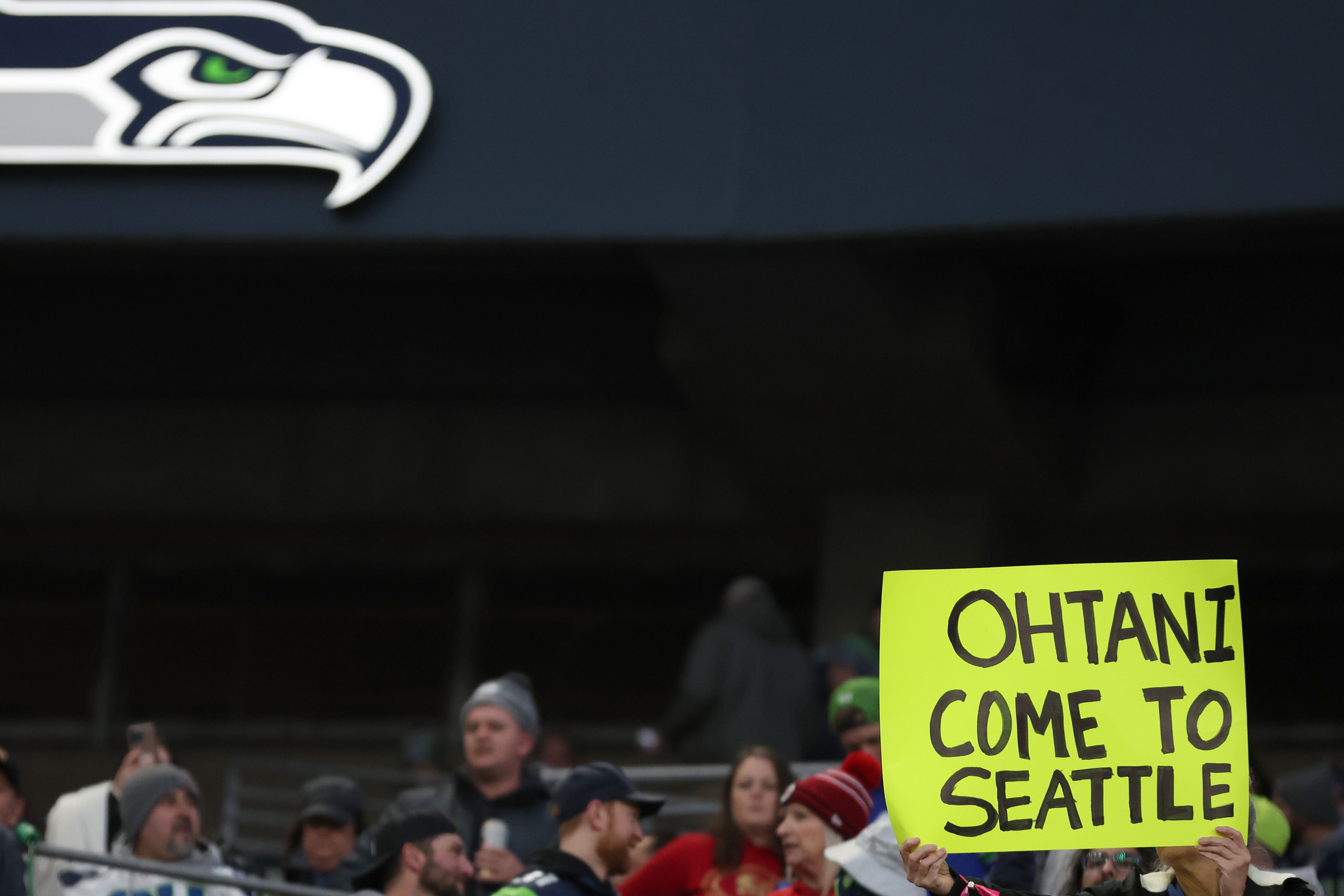 A fan holds a sign that reads "Ohtani come to Seattle" in a crowd below a Seahawks logo.