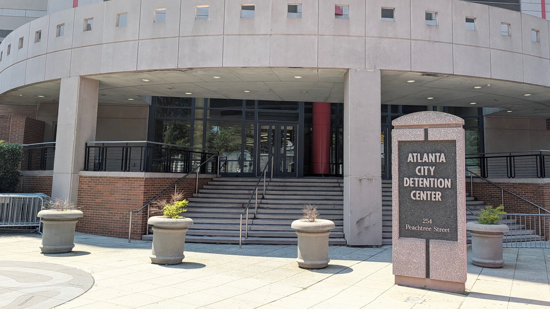 Exterior of the Atlanta City Detention Center: curved gray concrete facade with small square windows, glass doors, a red column, and stairs with railings; granite sign reads ATLANTA CITY DETENTION CENTER.