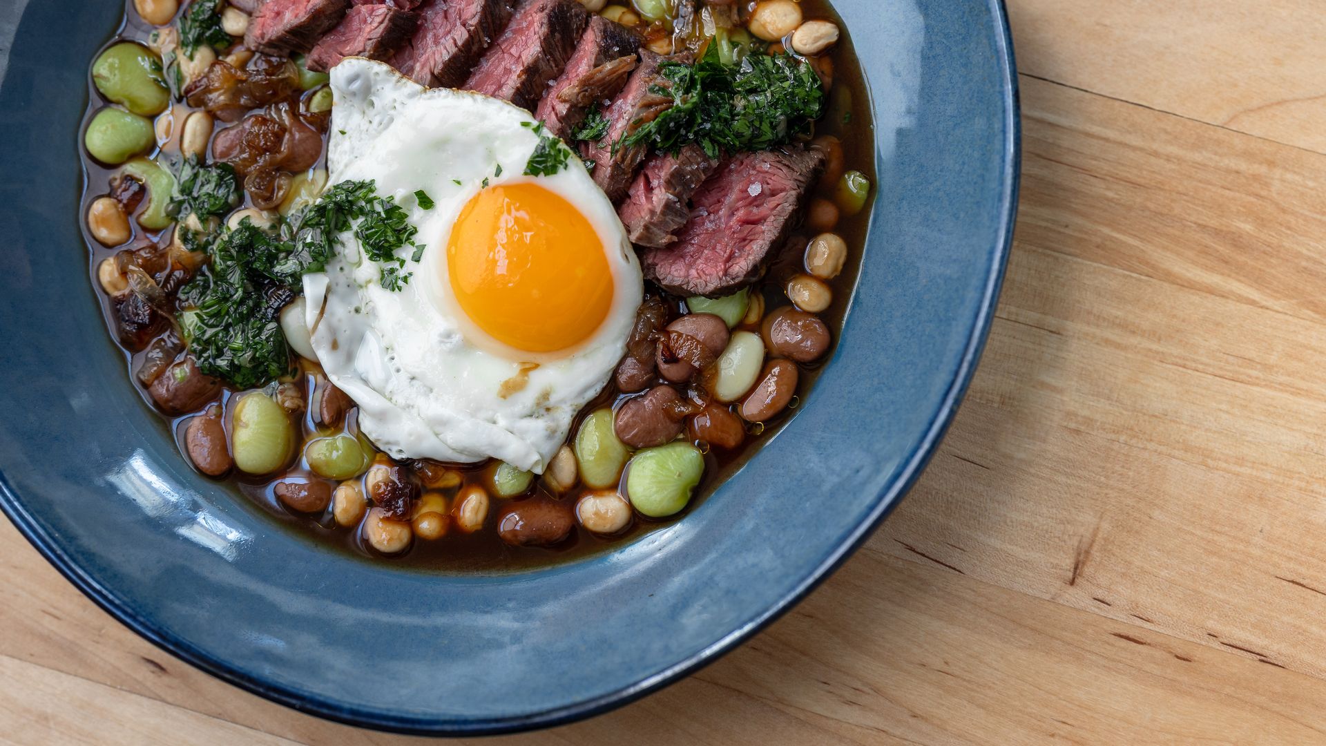 Blue bowl with sliced medium-rare steak, a fried egg with a bright yellow yolk, mixed beans, sauce, and green herb garnish on a wooden surface.