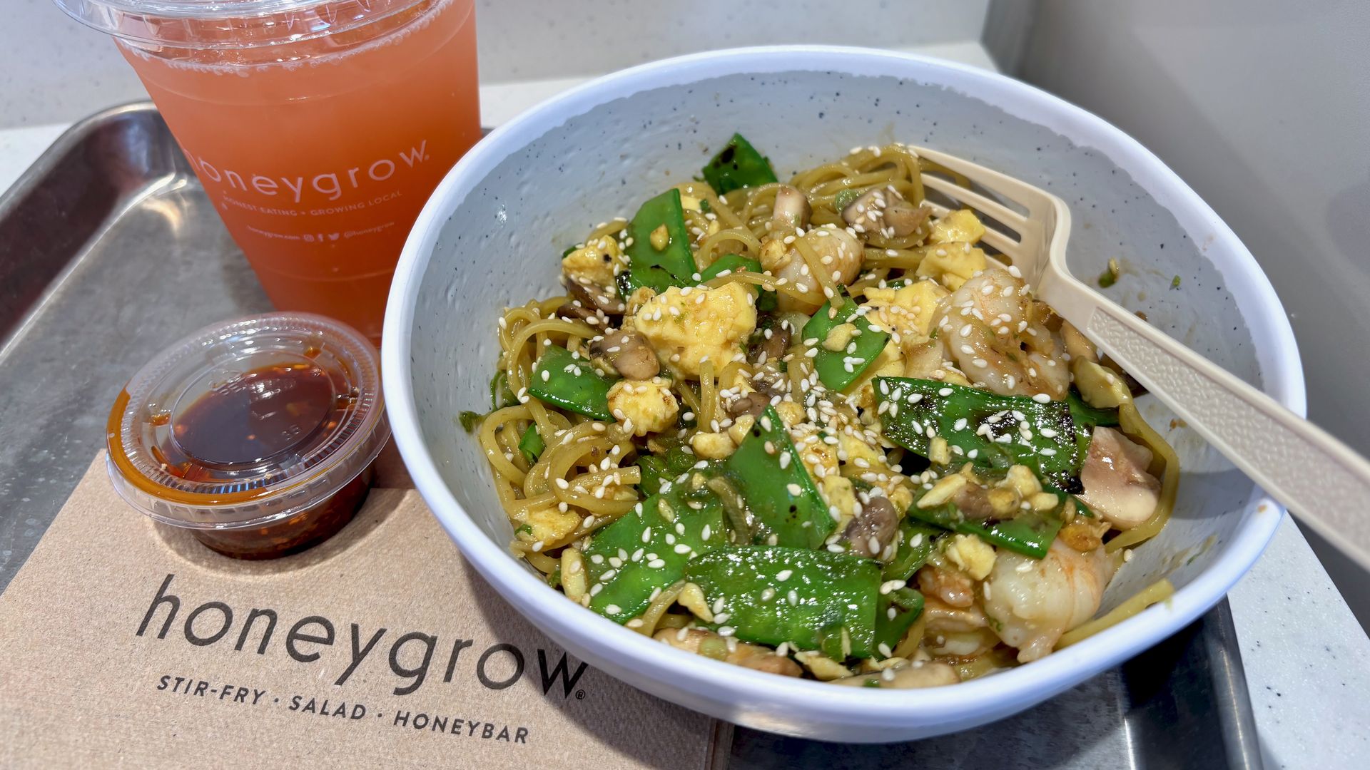 Bowl of stir-fry noodles with snow peas, mushrooms, shrimp, scrambled eggs, and sesame seeds, next to a small chili sauce container and a pink iced drink with a straw on a metal tray.