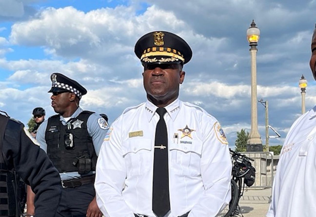 Black police captain in a white uniform, black tie, and peaked cap with gold insignia stands outdoors under a cloudy blue sky; background shows officers in tactical vests and decorative streetlamps.