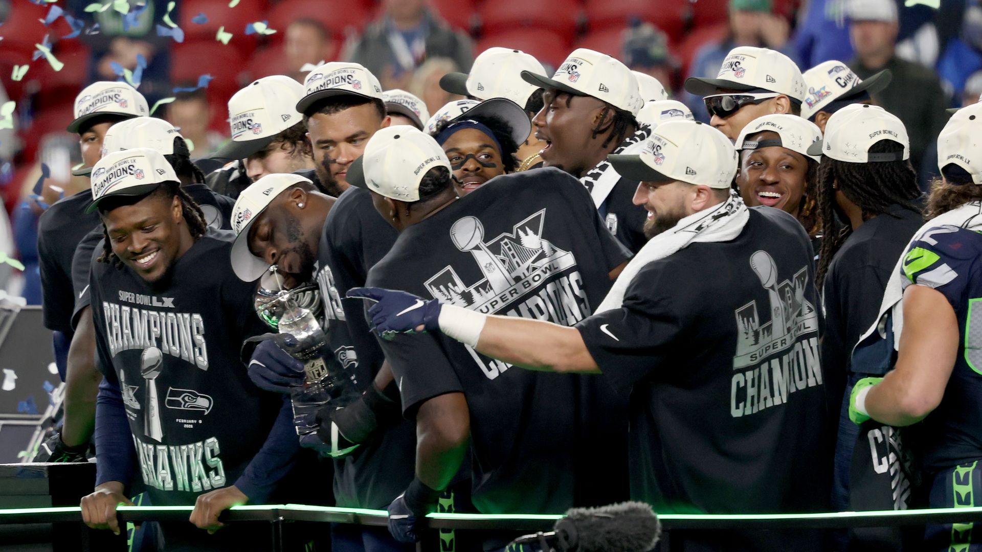 Members of the Seattle Seahawks team gather while wearing ball caps and shirts that say Champions as they celebrate with ticker tape falling around them.