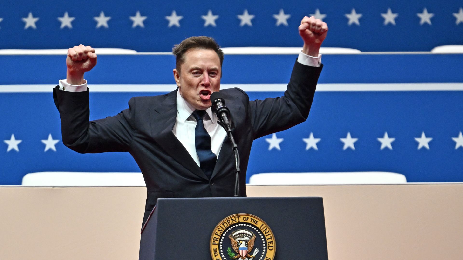 Elon Musk wearing a gray suit and holding up both of his fists while standing at a podium in front of a blue starred backdrop.