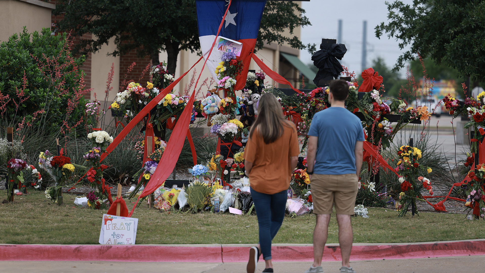 Two people look at a memorial to those killed at the Allen Premium Outlets mall...lots of flowers and ribbon and a Texas flag