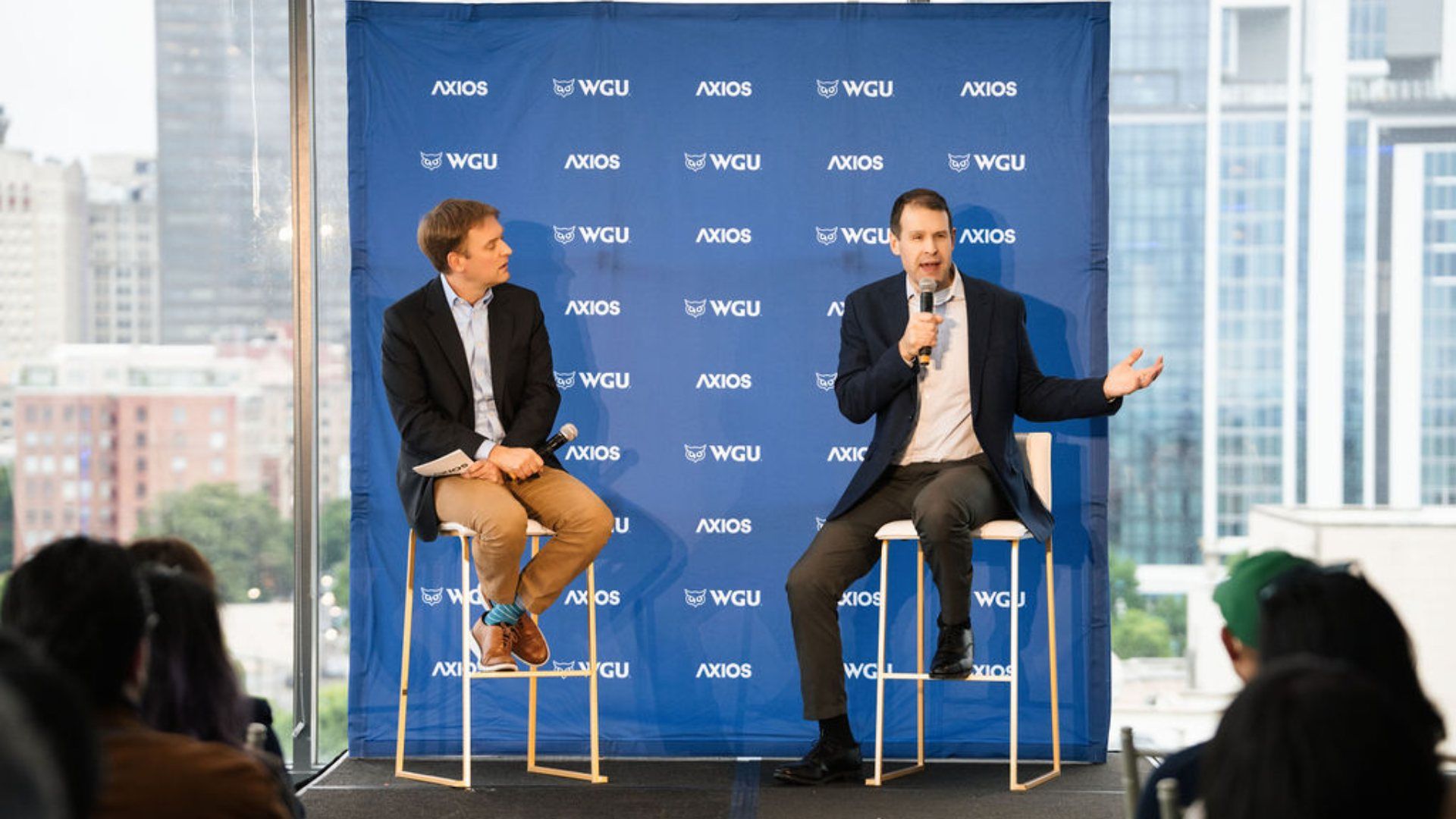 Two men sit on high stools on a stage with a blue backdrop featuring the logos "WGU" and "AXIOS"; one speaks into a microphone and gestures, while the other watches, with a city skyline in the background.