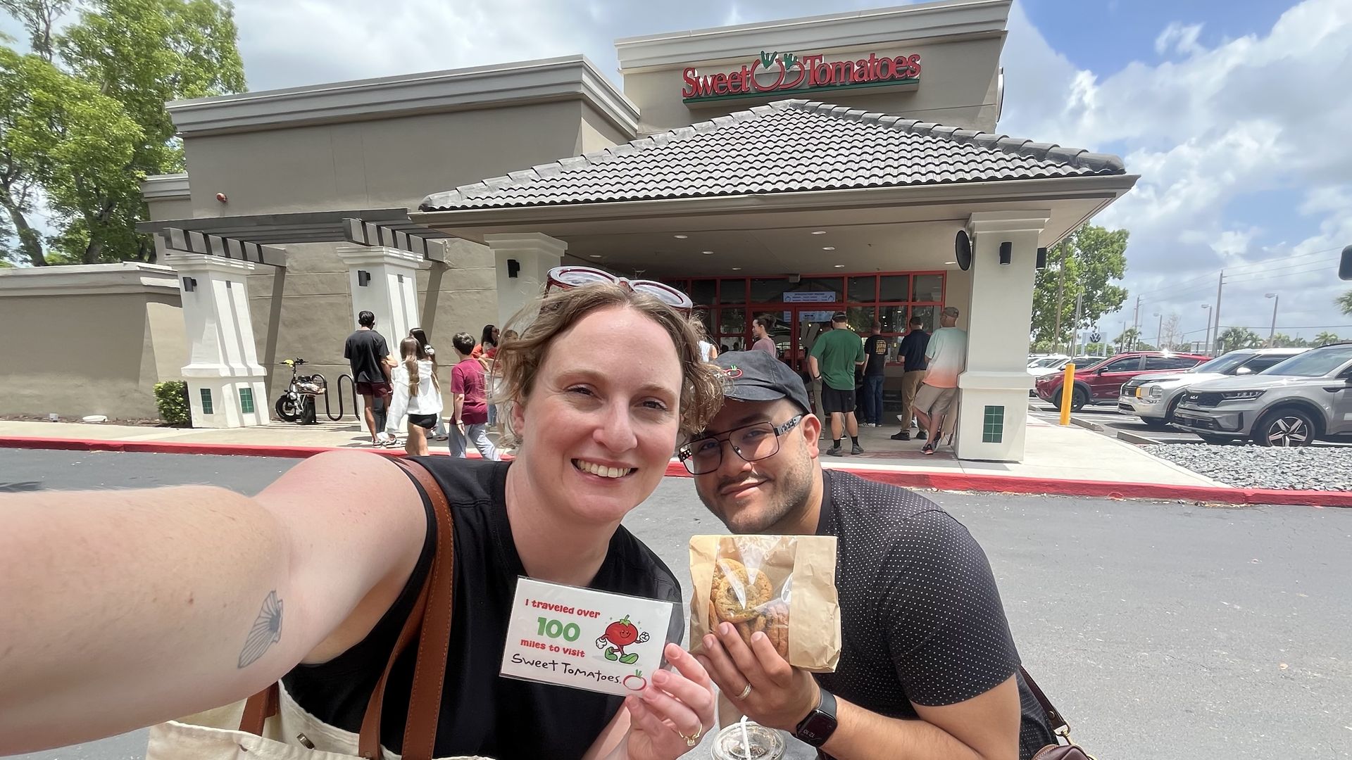 Smiling friend selfie outside Sweet Tomatoes, holding a bag and a card. A line forms at the entrance; sunny day, blue sky, green trees, and parked cars in the lot.