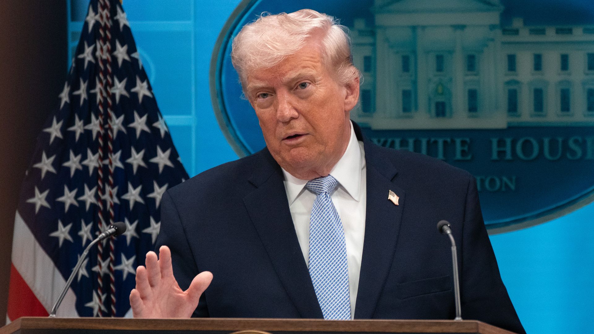 President Donald Trump speaks at a podium with the presidential seal, wearing a dark suit and light blue tie, flanked by American flags, against a blue backdrop with the White House emblem.