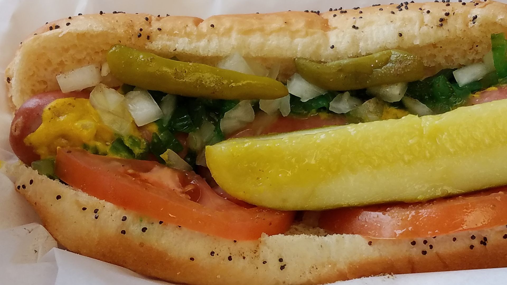 Photo of a hot dog with all of the ingredients. Onions, tomatoes, pickles and sport peppers in a bun. 