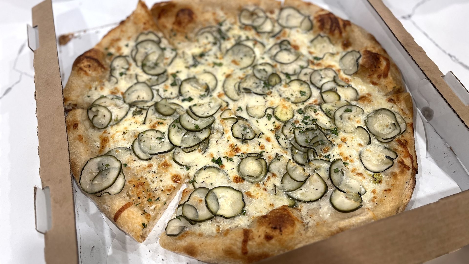 A whole pizza with pickles on top in a pizza box.