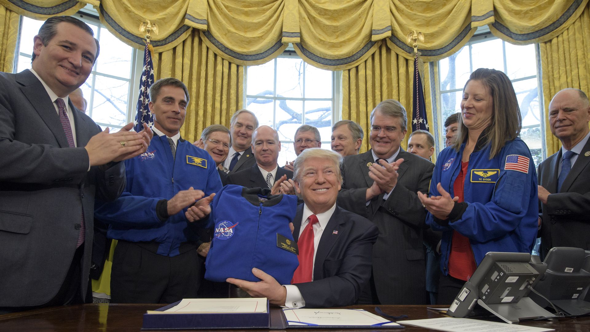 President Trump sits behind a desk, surrounded by NASA staff and Ted Cruz. Trump is holding a NASA launch jacket that he has just been gifted.