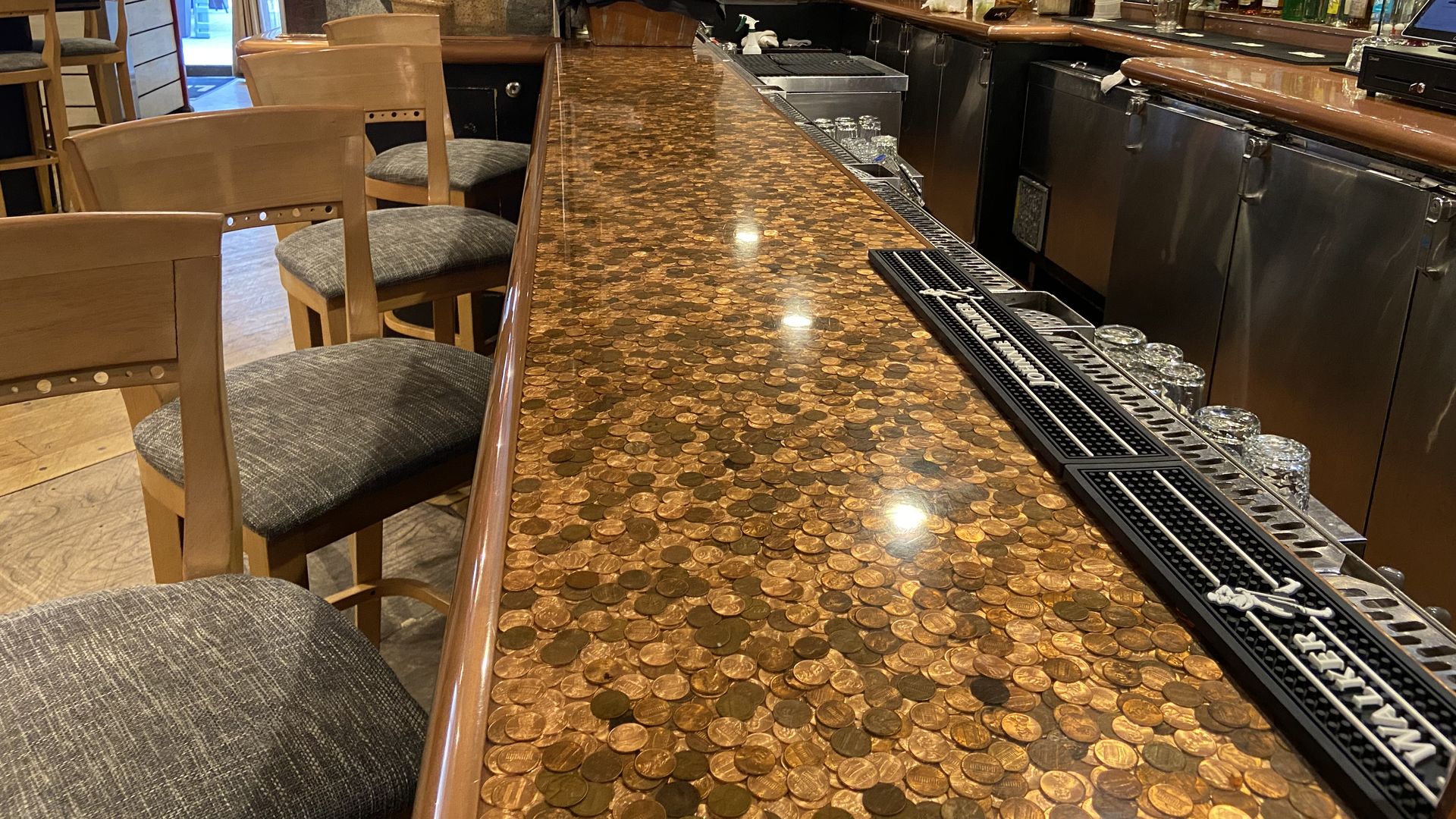 Empty bar with a countertop covered in pennies, surrounded by wooden chairs with gray cushions, shelves stocked with bottles, a TV, and an entrance with red framing in the background.