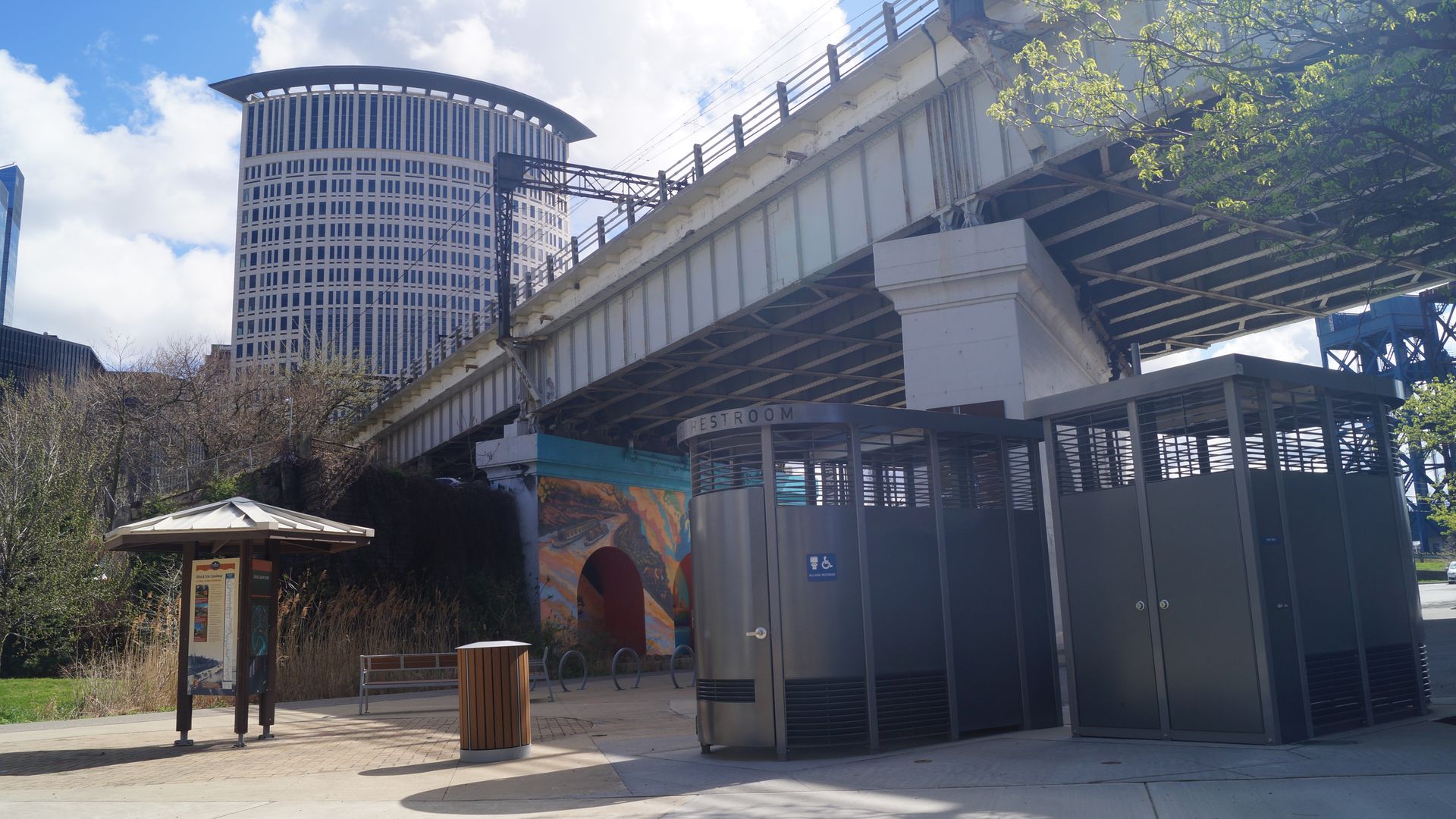 Urban scene with a white elevated roadway spanning the image, a curved cylindrical high-rise on the left, blue sky and clouds, green trees on the right, and a restroom entrance in the foreground.