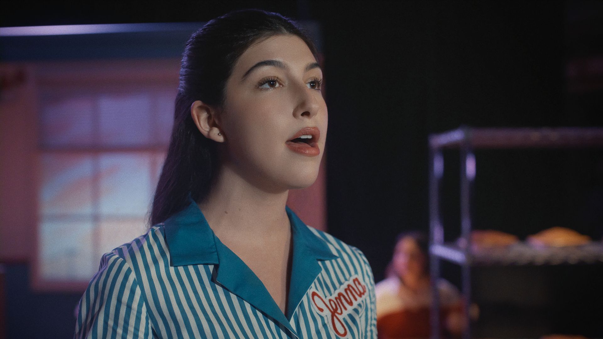 Close-up of a young woman with dark hair wearing a blue and white striped shirt, looking upward with a thoughtful expression; purple lighting and a blurred audience in the dark background.