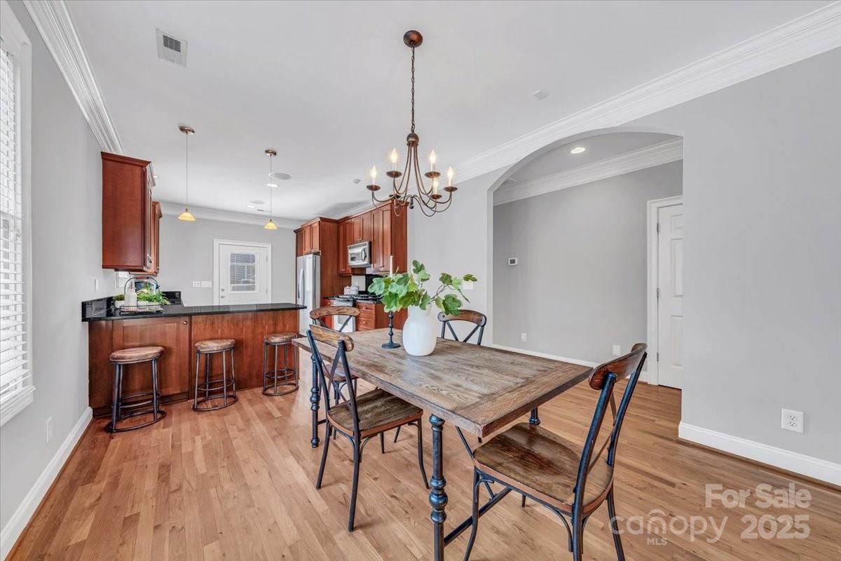 Bright kitchen dining area with wooden floors, wood cabinets, black countertops, a wooden table with four metal-framed chairs, green plant centerpiece, and chandelier lighting.