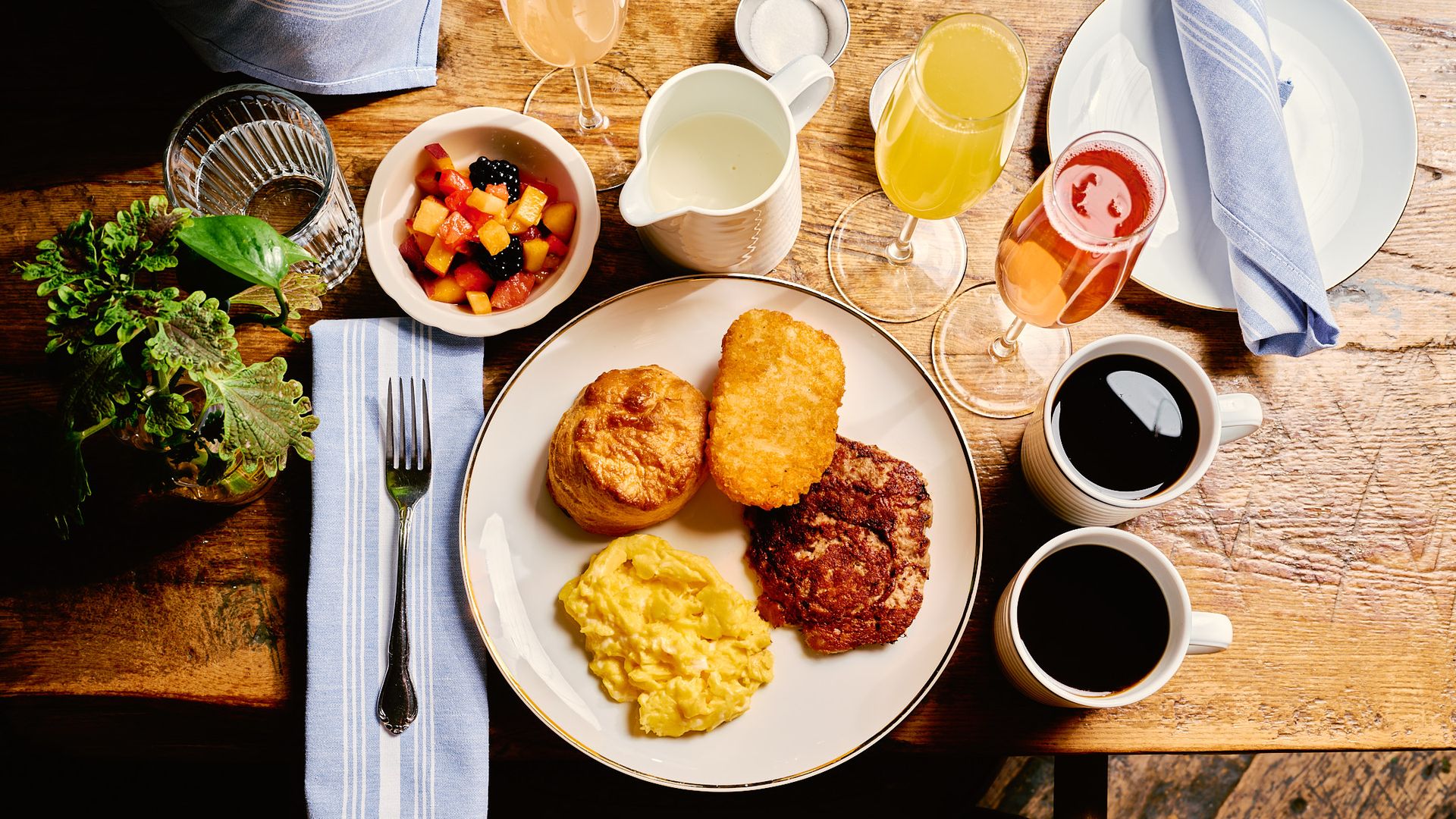 Breakfast plate with scrambled eggs, sausage patty, hash brown, and biscuit on wooden table with fruit bowl, two cups of black coffee, two glasses of orange and red juice, blue napkin, and fork.
