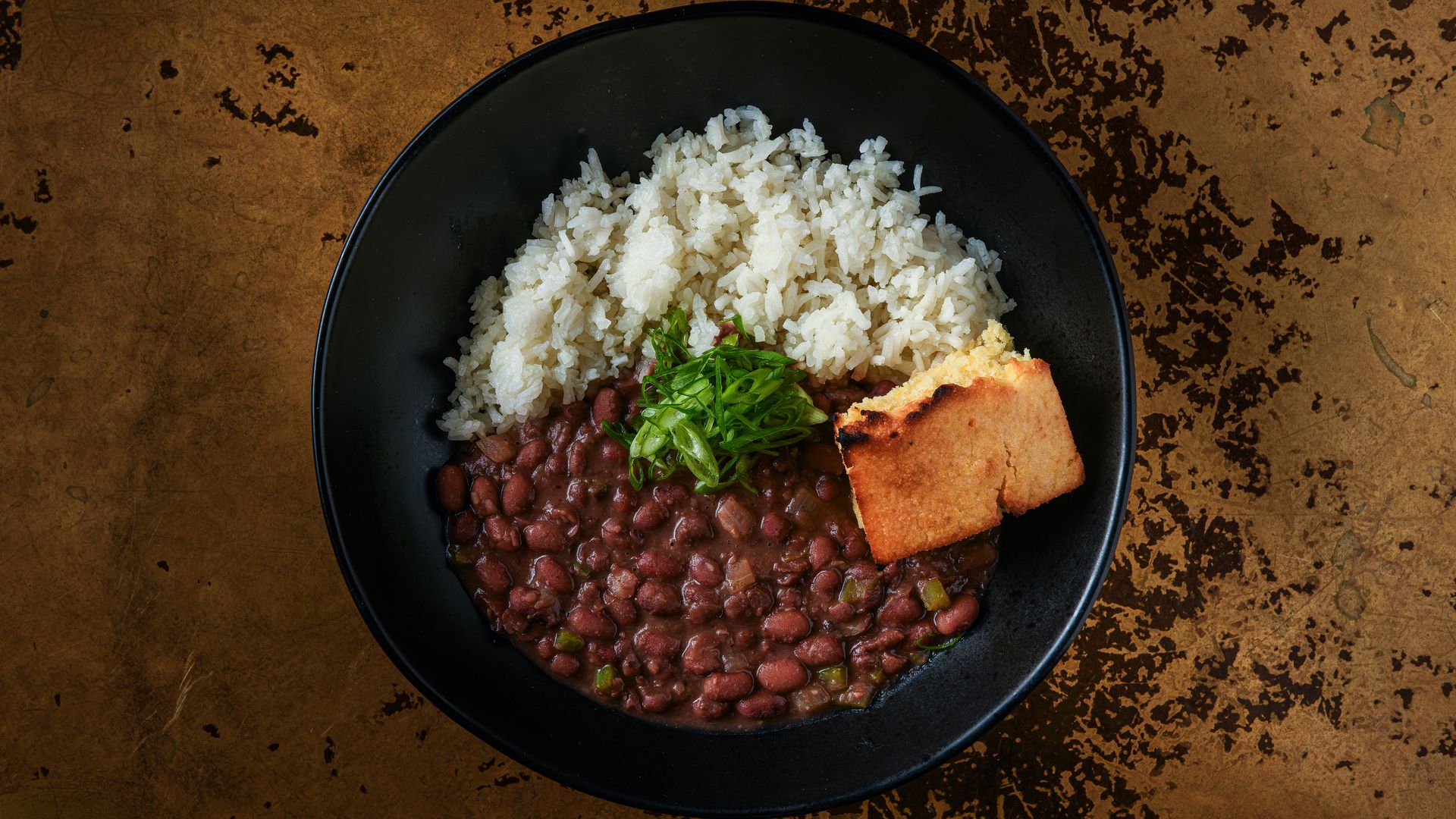 Black bowl with white rice, brown beans in sauce topped with green onions, and a golden-brown cornbread piece on a textured brown surface.