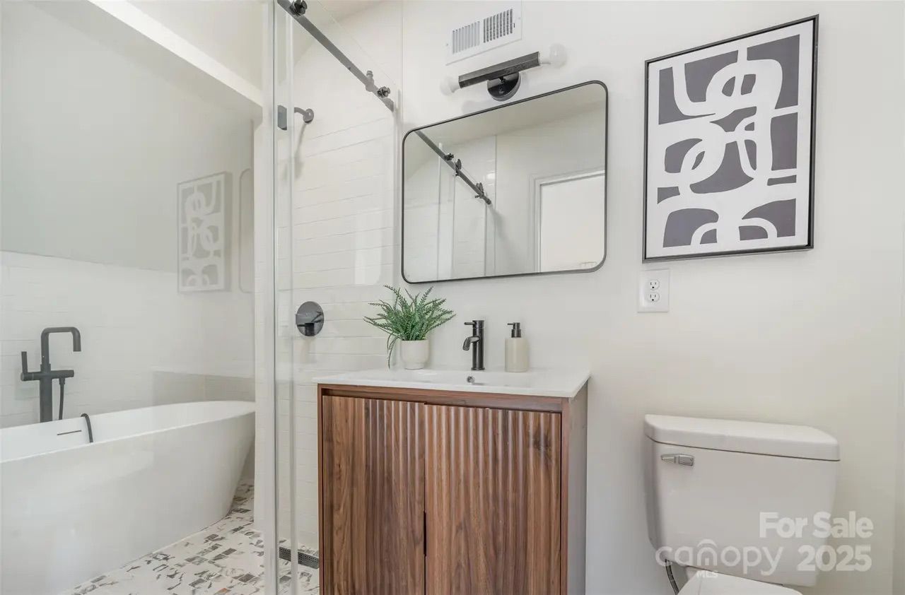 Modern bathroom with white bathtub, glass shower door, wooden vanity with black faucet, large rectangular mirror, abstract black and white wall art, and white toilet on patterned tile floor.