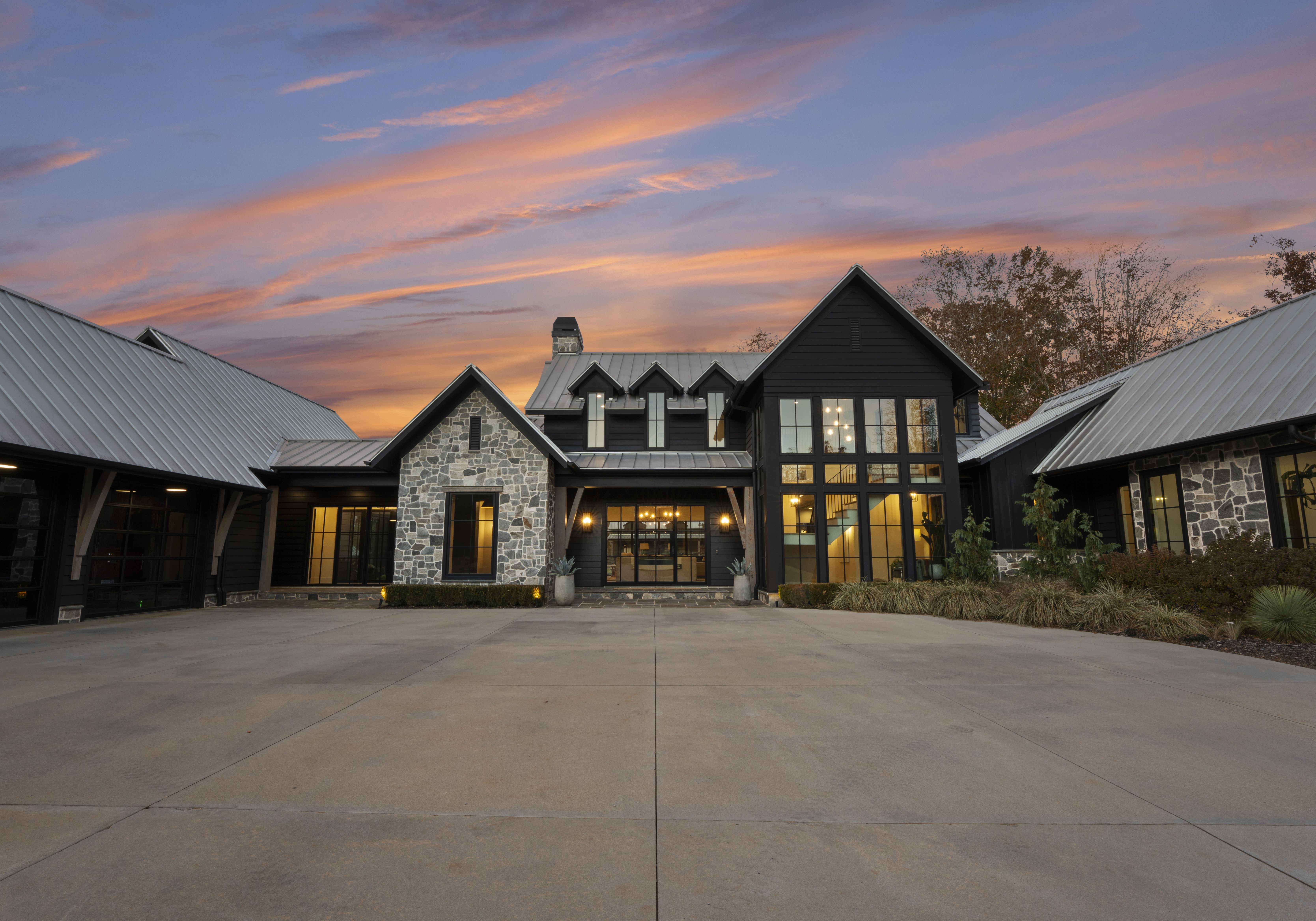 Front view of a modern stone and dark wood building with metal roofs, large glass windows, and warm interior lights, set against a pink-orange sunset; wide concrete driveway in foreground.