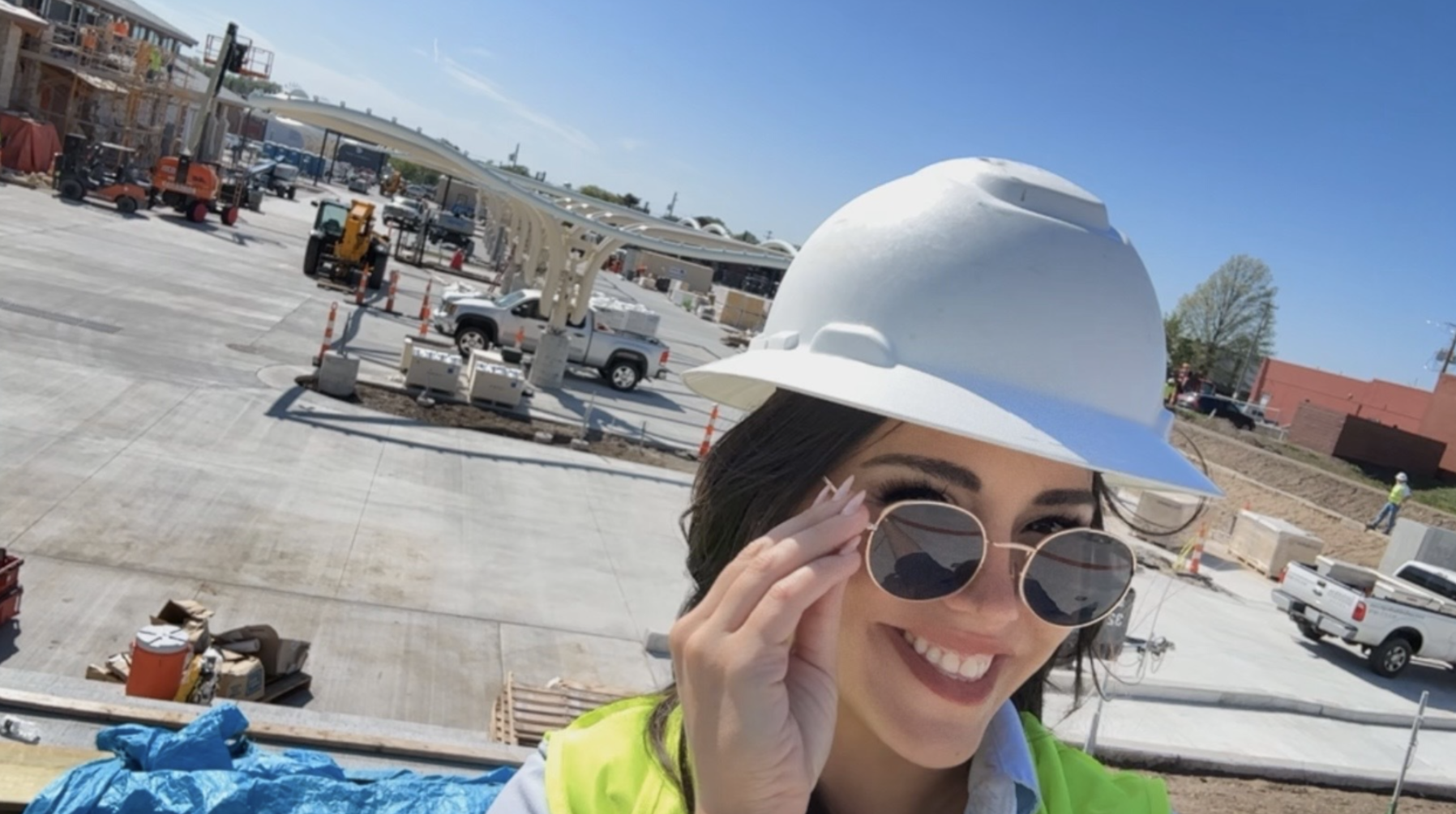 Smiling woman in a white hard hat and round sunglasses selfies at a sunny construction site with orange machinery, trucks, and a white curved structure; wearing a neon safety vest.