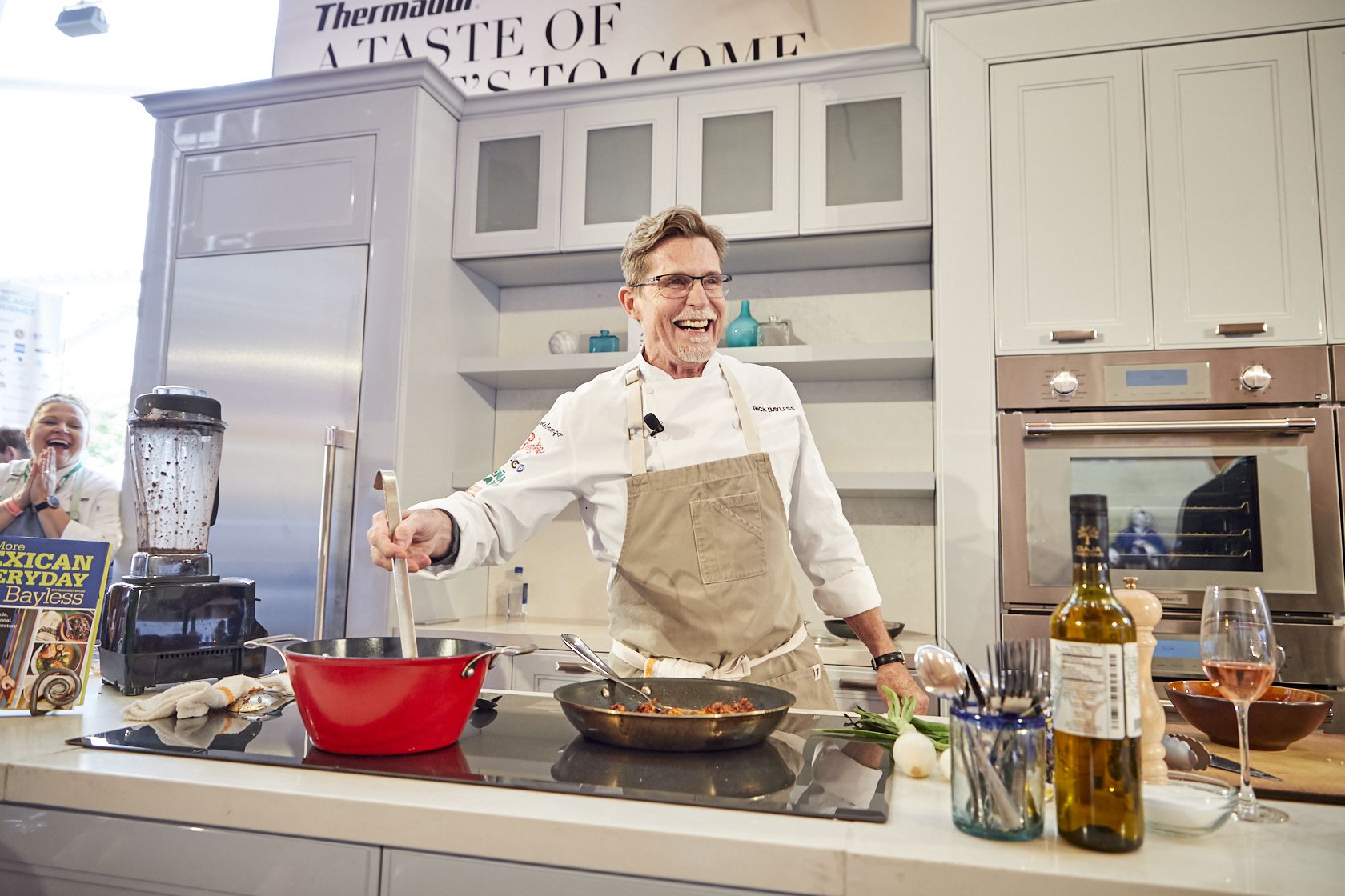 Chef in beige apron and white shirt demonstrates cooking in a kitchen studio, smiling while stirring a skillet. Blender, red pot, wine bottle, and glass on counter; cheering spectator in background.