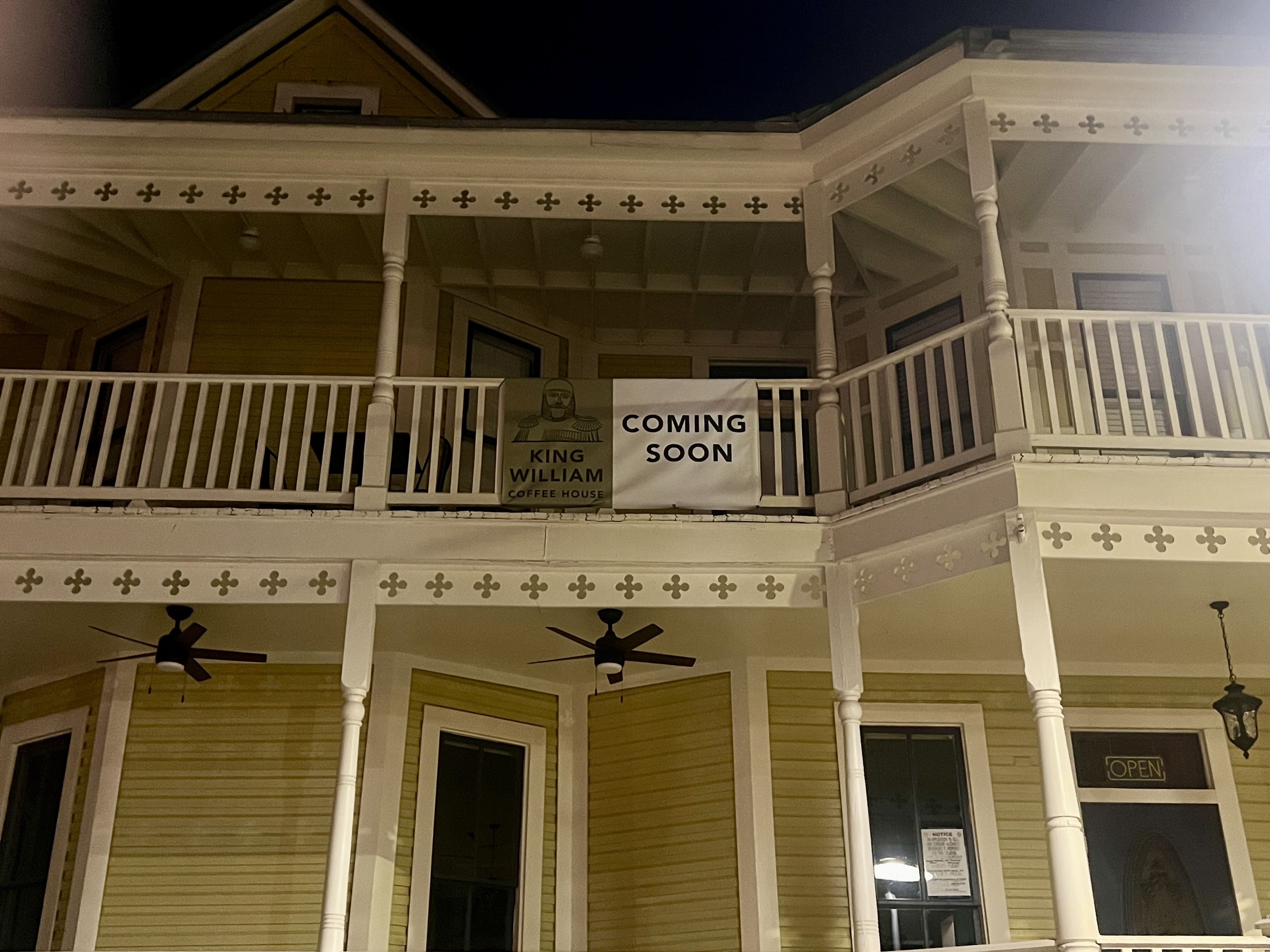 Yellow two-story building with white trim and railings at night. Signs on second-floor balcony read "King William Coffee House" and "Coming Soon." A door below displays an "Open" sign.