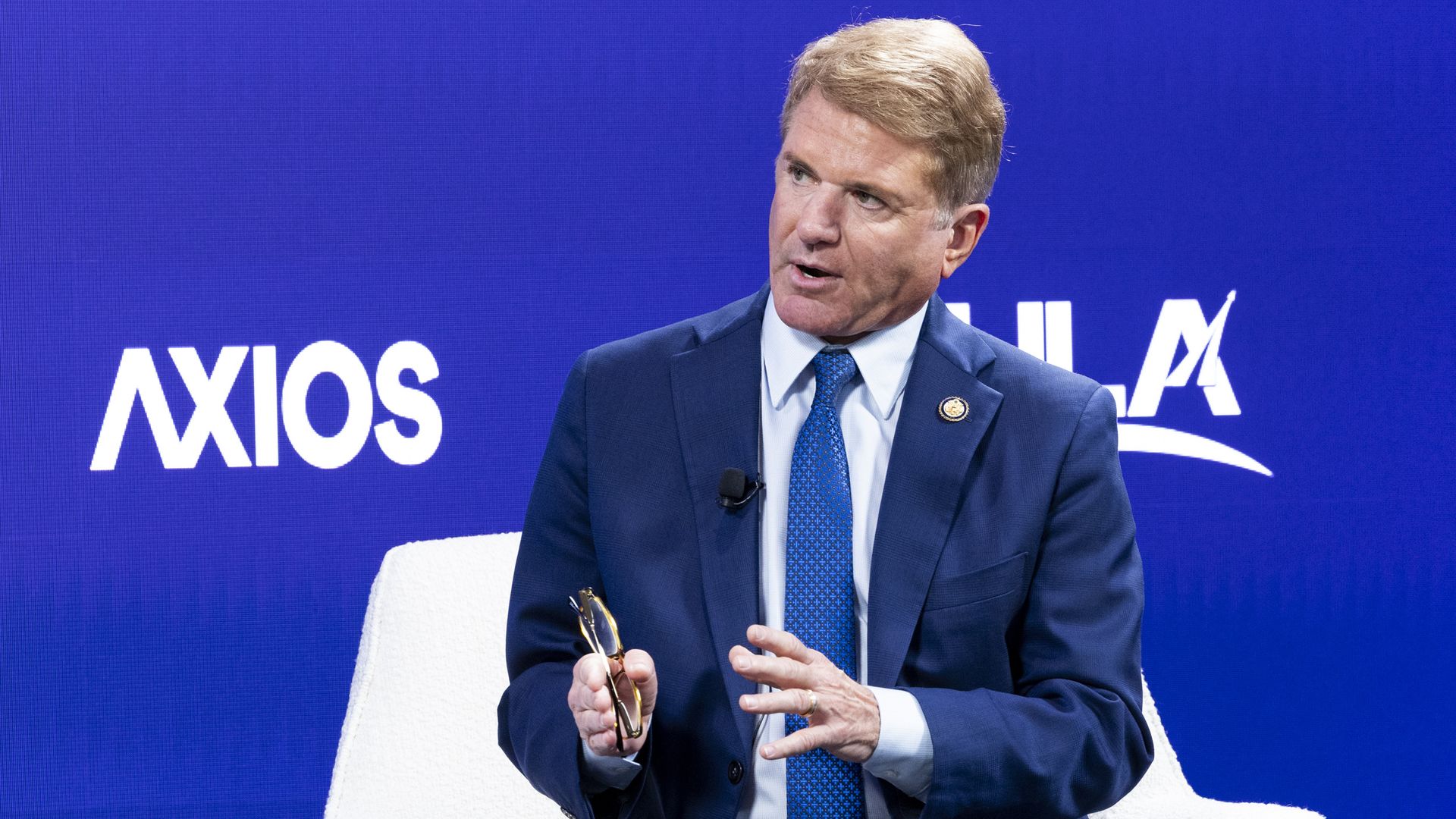 Man in a blue suit, light blue shirt, and blue patterned tie speaking while seated on a white chair against a blue backdrop with Axios logo.
