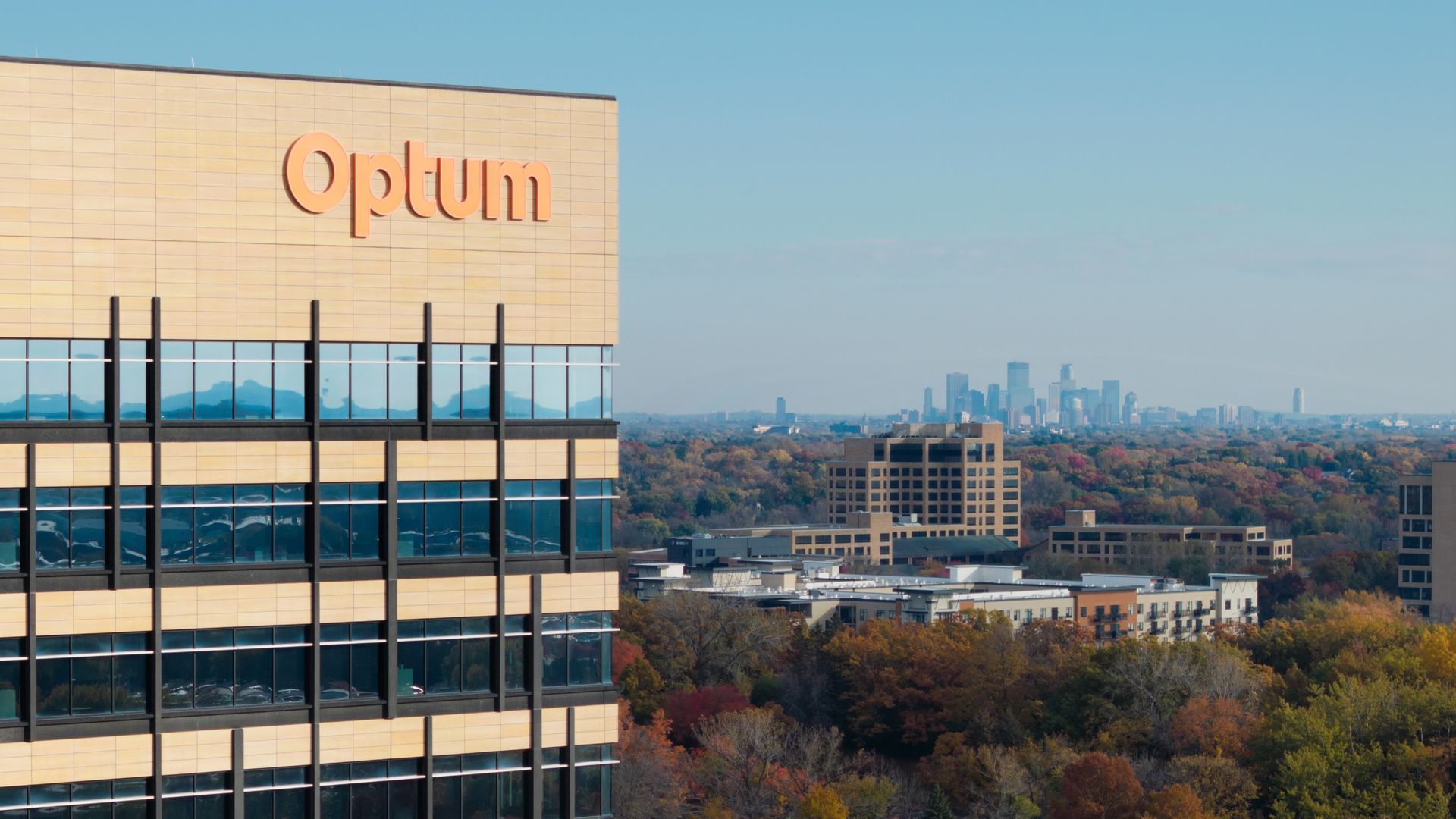 Modern office building with beige panels and glass windows, featuring an orange Optum sign. Distant city skyline under a clear blue sky, with autumn trees and a road in the foreground.
