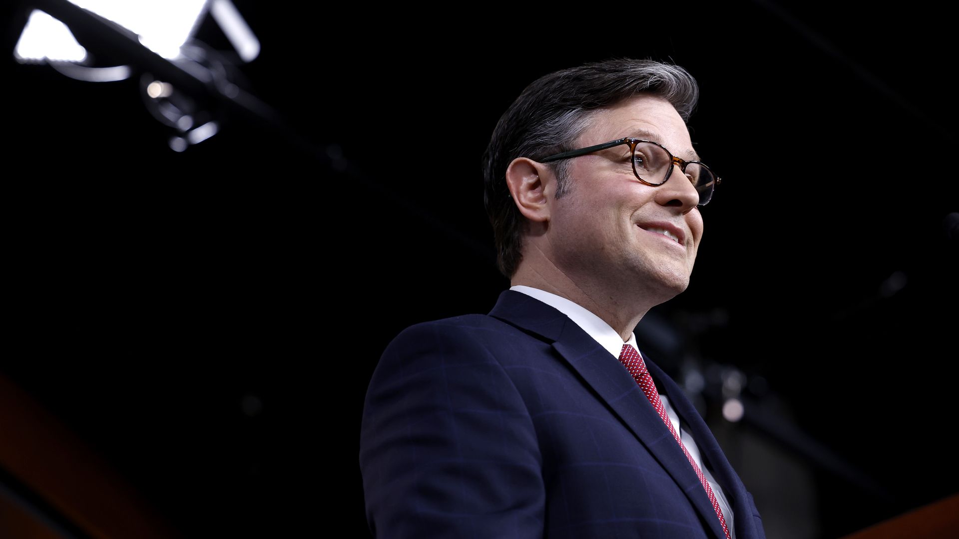 House Speaker Mike Johnson, wearing a blue suit, white shirt, red tie and glasses in a TV studio at the Capitol.