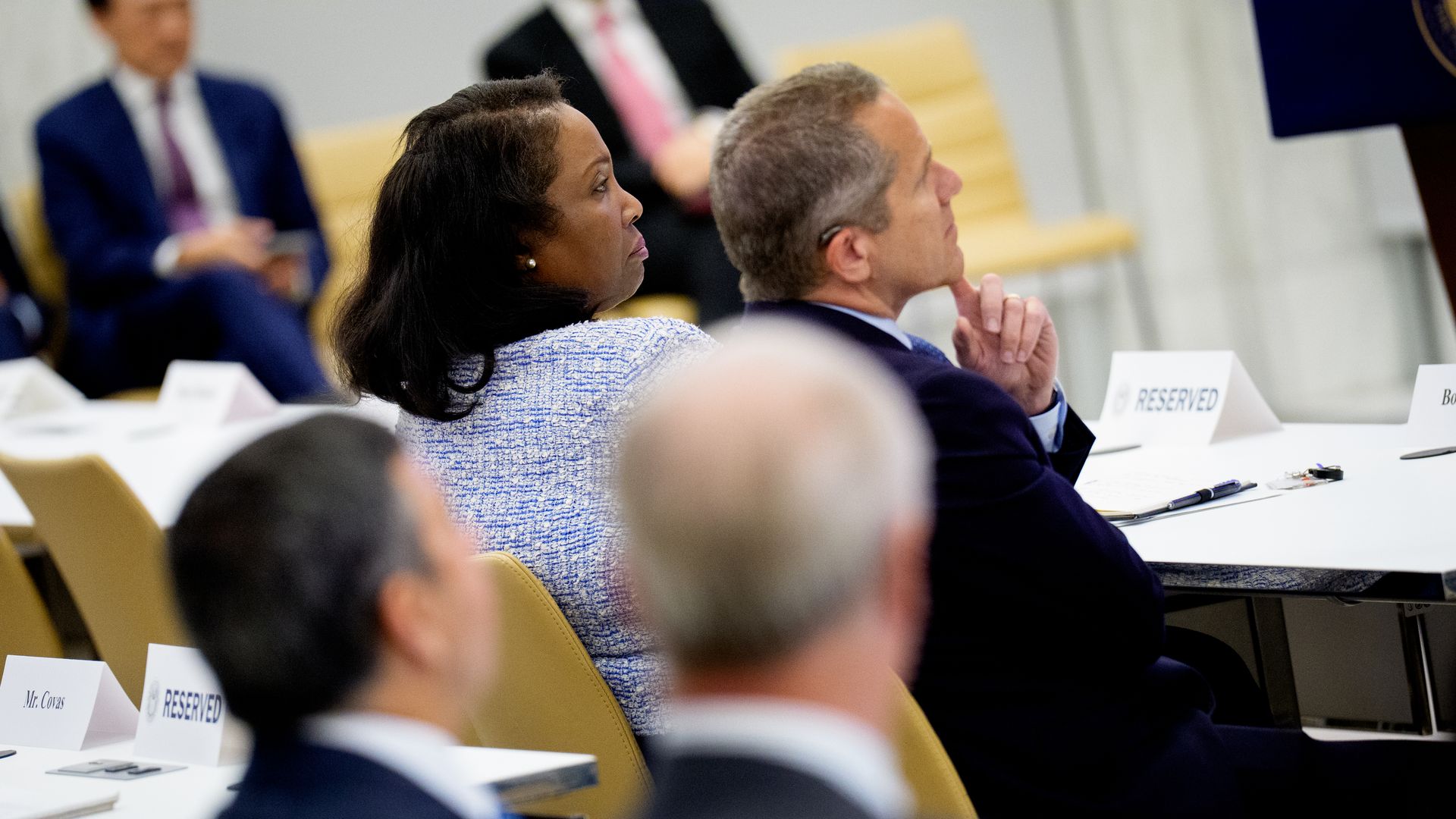 People in business attire seated attentively in a meeting room with signs reading "RESERVED" on tables, cream-colored chairs, and notebooks and pens on the tables.