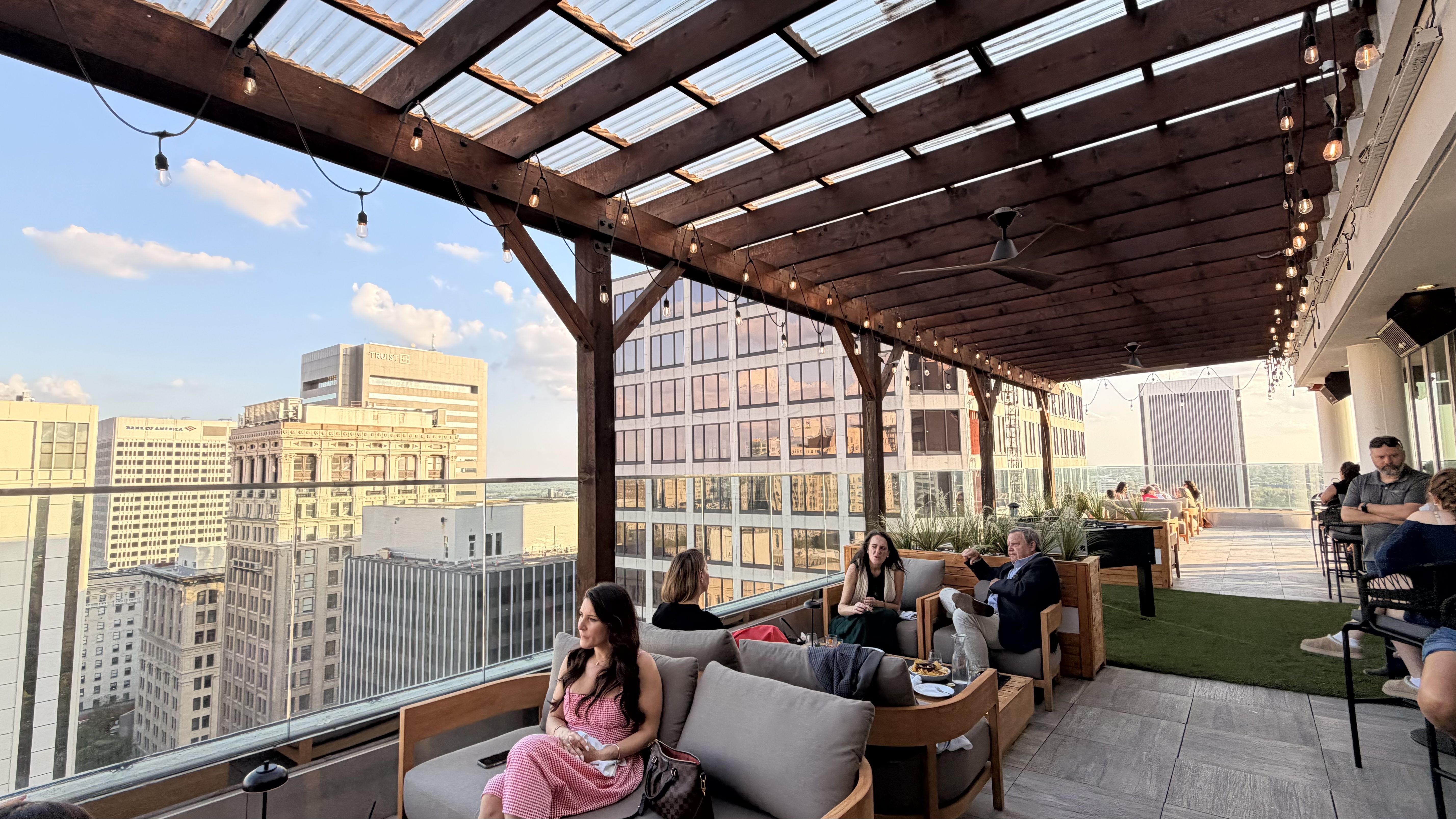 Rooftop lounge with a wooden pergola and string lights, overlooking a city skyline. People relax on cushioned seating, chatting and dining as the sunset glows near the glass railing.
