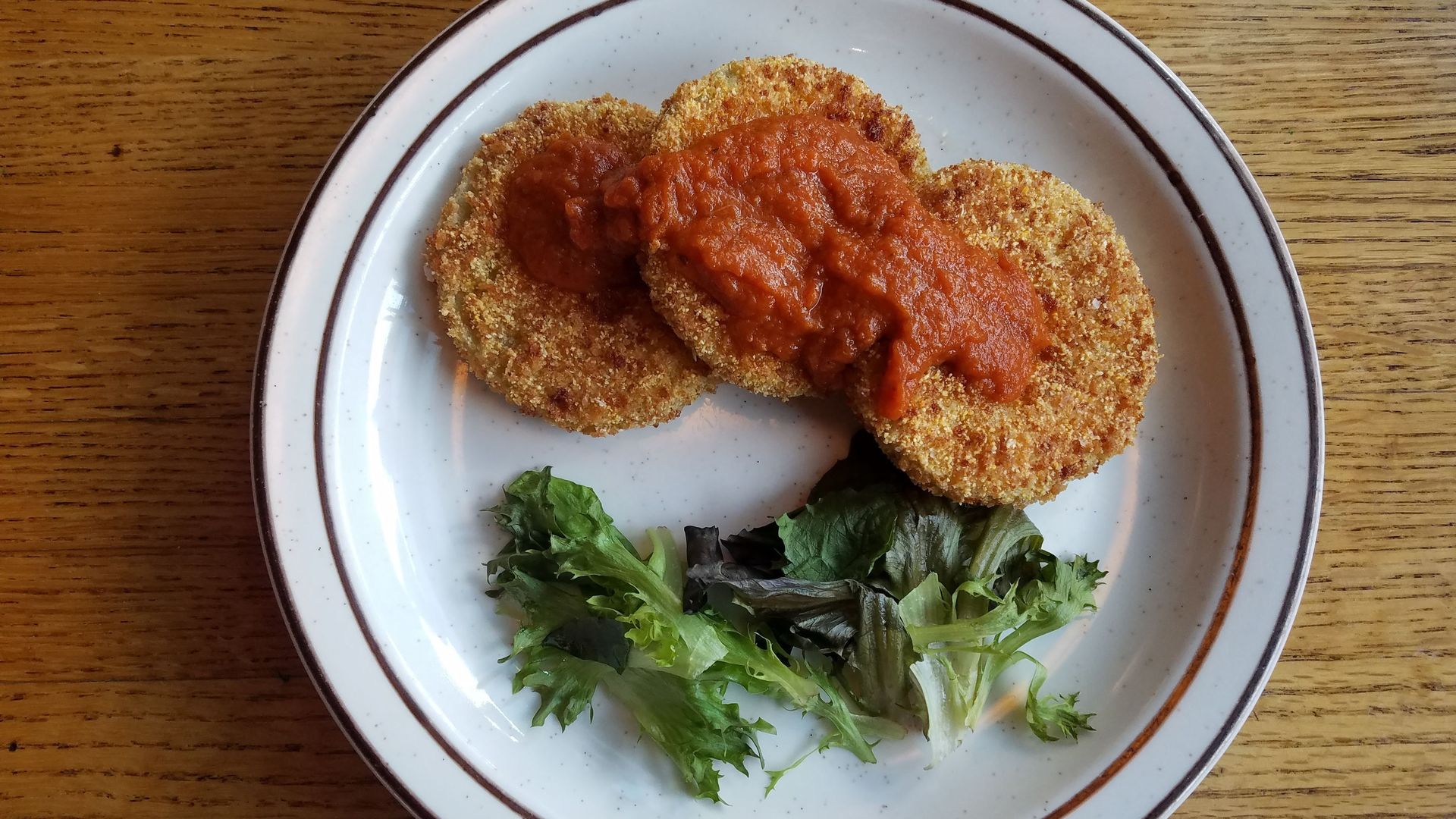 Two breaded fried croquettes topped with red tomato sauce on a white plate with a brown rim, set on a wooden table, with leafy greens on the side.
