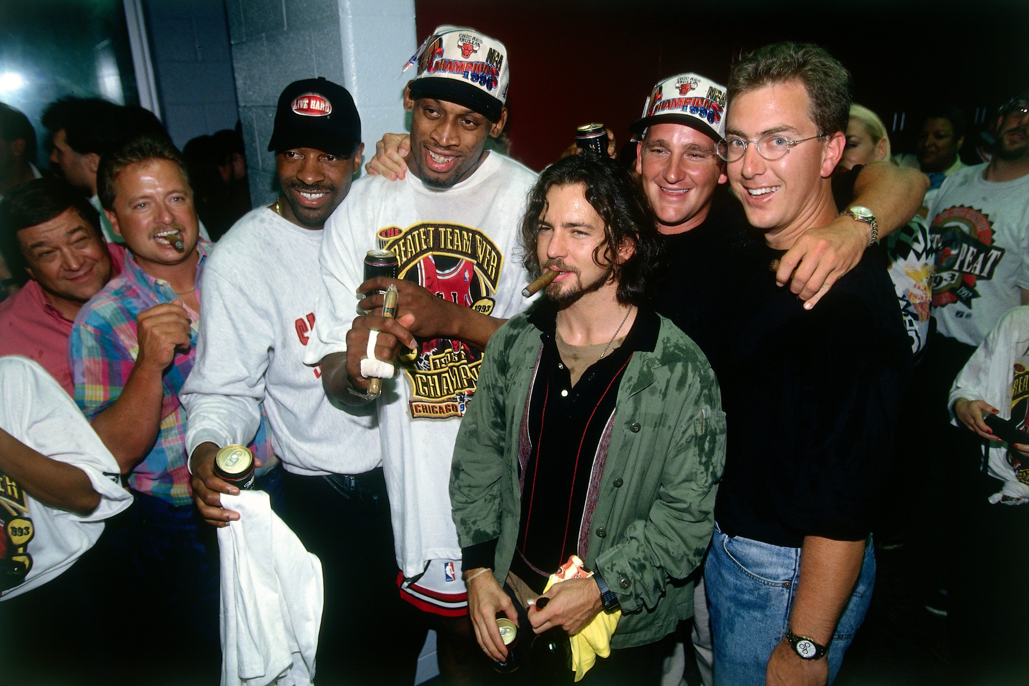 Group of men at a celebration in a hallway, some wearing white Bulls championship hats; they smile, hold drinks or cigars, and hug or stand close with arms around each other.