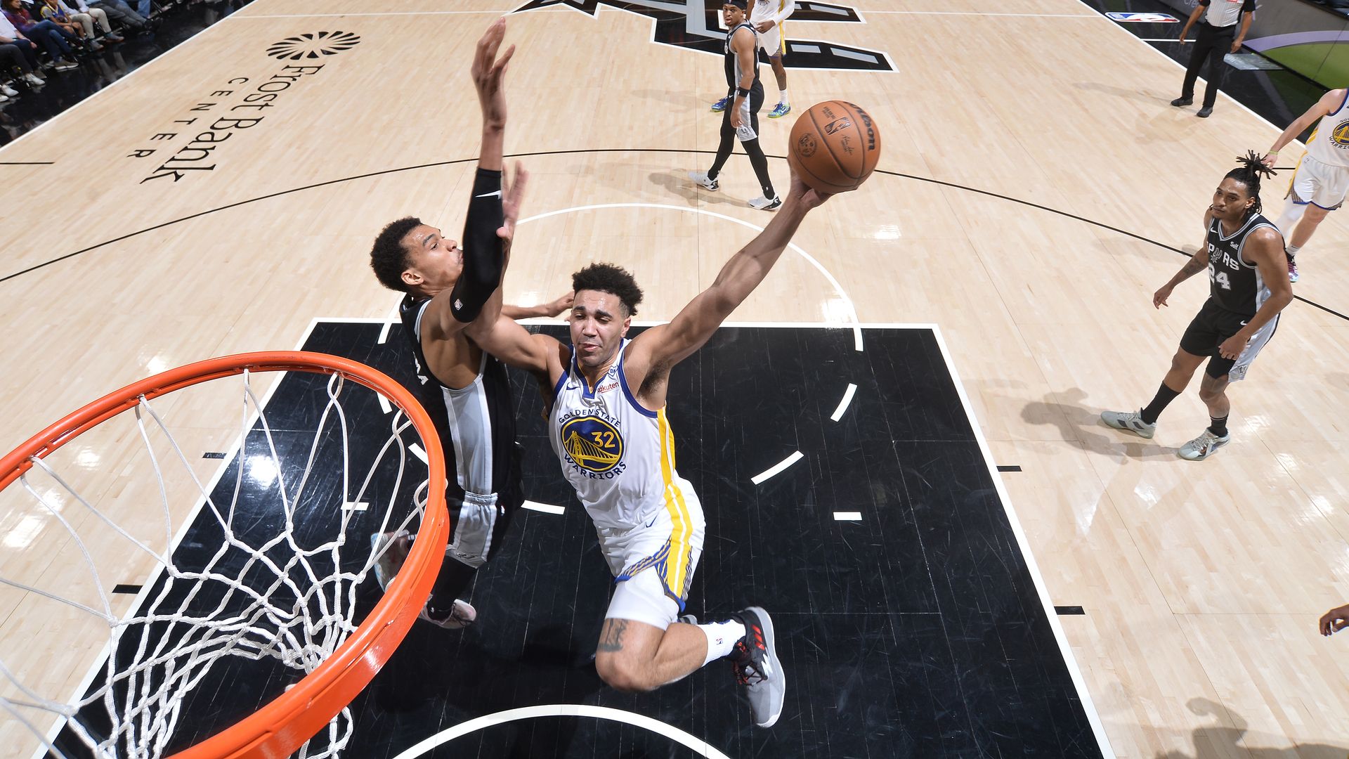 a basketball player in a white warriors jersey drives toward the basket, challenging a player wearing black at the rim