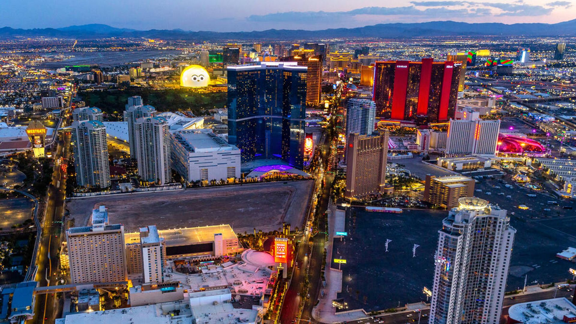 Aerial view of Las Vegas Strip at dusk with illuminated hotels and neon signs; blue and red towers line the skyline, busy roads below, and a large glowing yellow emoji face near the center.
