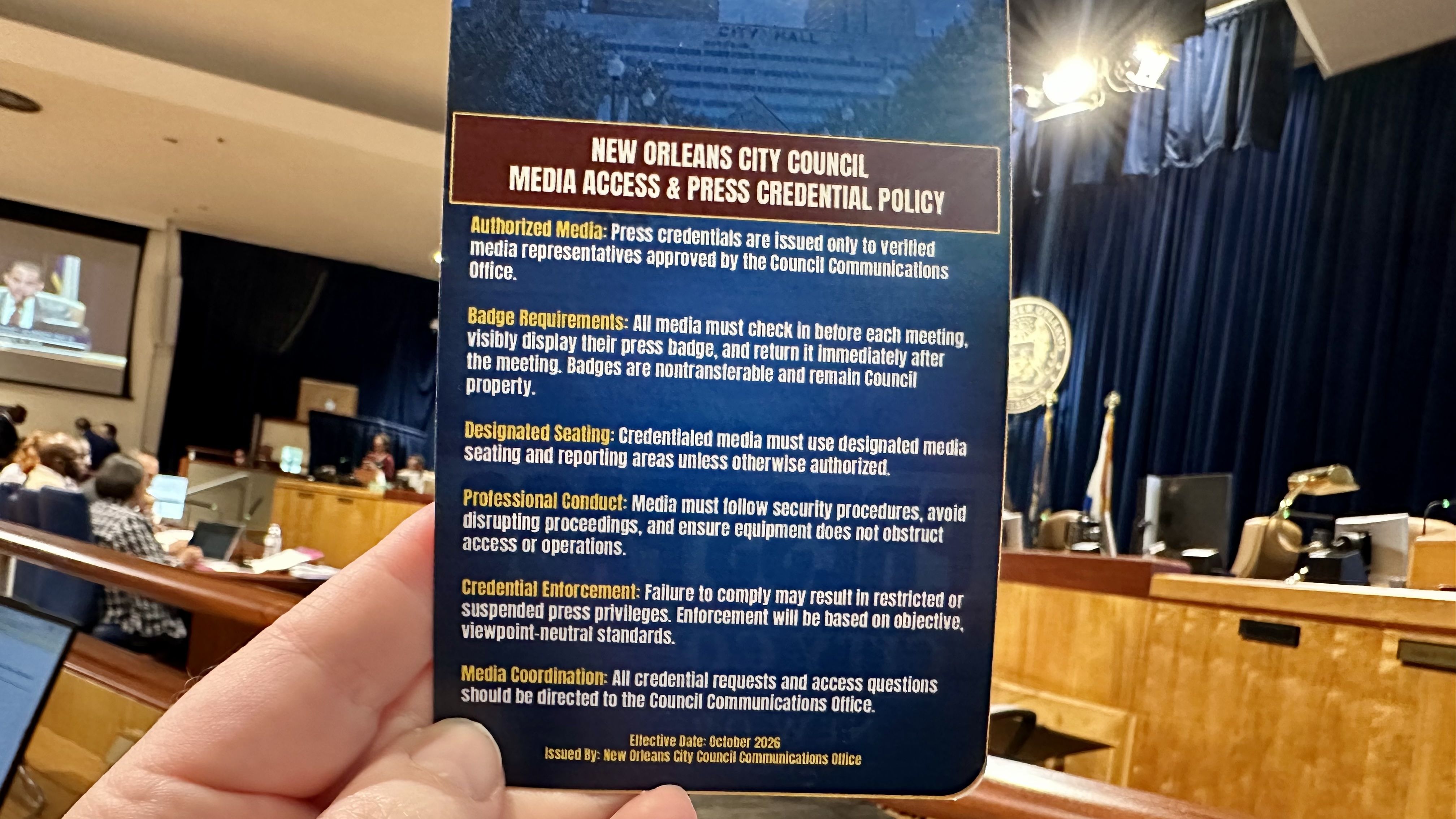 A hand holds a tall blue poster titled "New Orleans City Council: Media Access & Press Credential Policy" inside a busy council chamber with attendees, a large screen, and a seal on a blue curtain in the background.