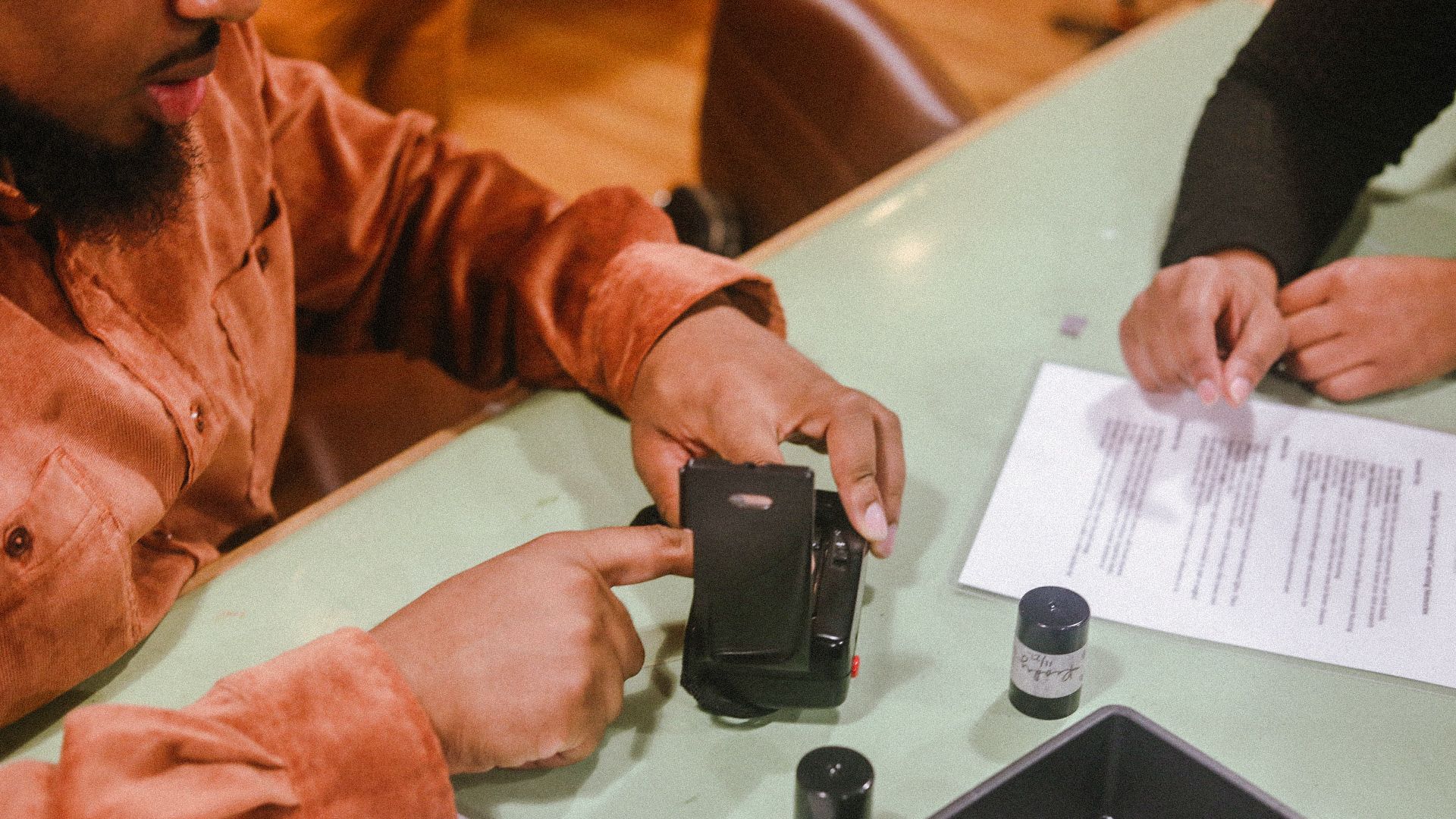Two people at a light green table examining a black instant camera, film canisters, scissors, and printed papers; one person wears an orange shirt, the other black.