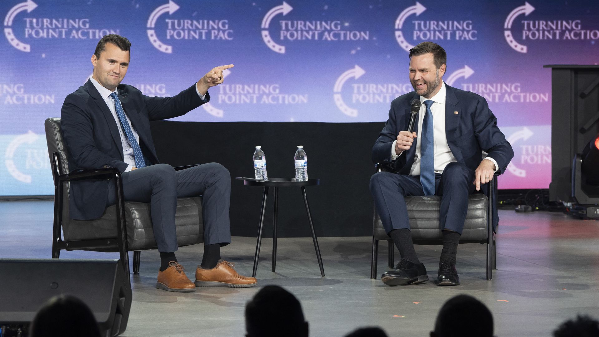 Two men in suits seated on stage during a Turning Point Action event, one holding a microphone and laughing, the other pointing, with water bottles on a small table between them.